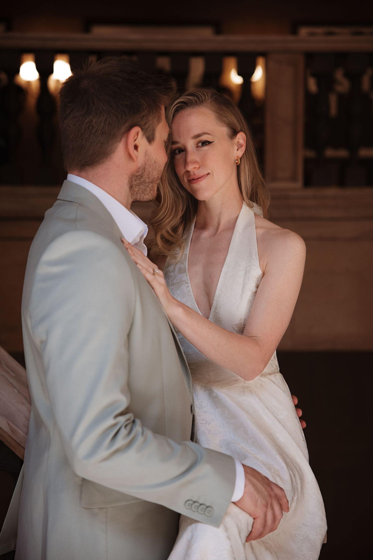 Bride in white halter gown gazes at groom in beige suit during romantic indoor wedding portrait.