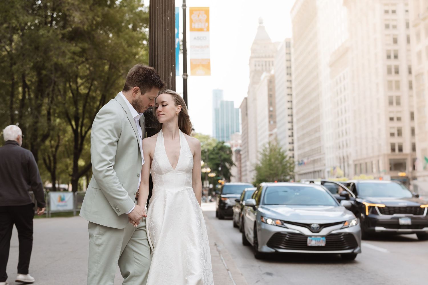 A bride and groom walk hand in hand on a sunny city street, smiling at each other in elegant wedding attire.