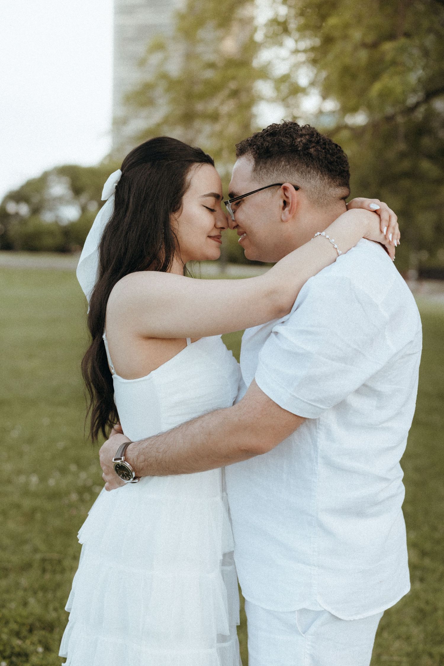 Romantic couple in white outfits touching foreheads in a park with city backdrop.