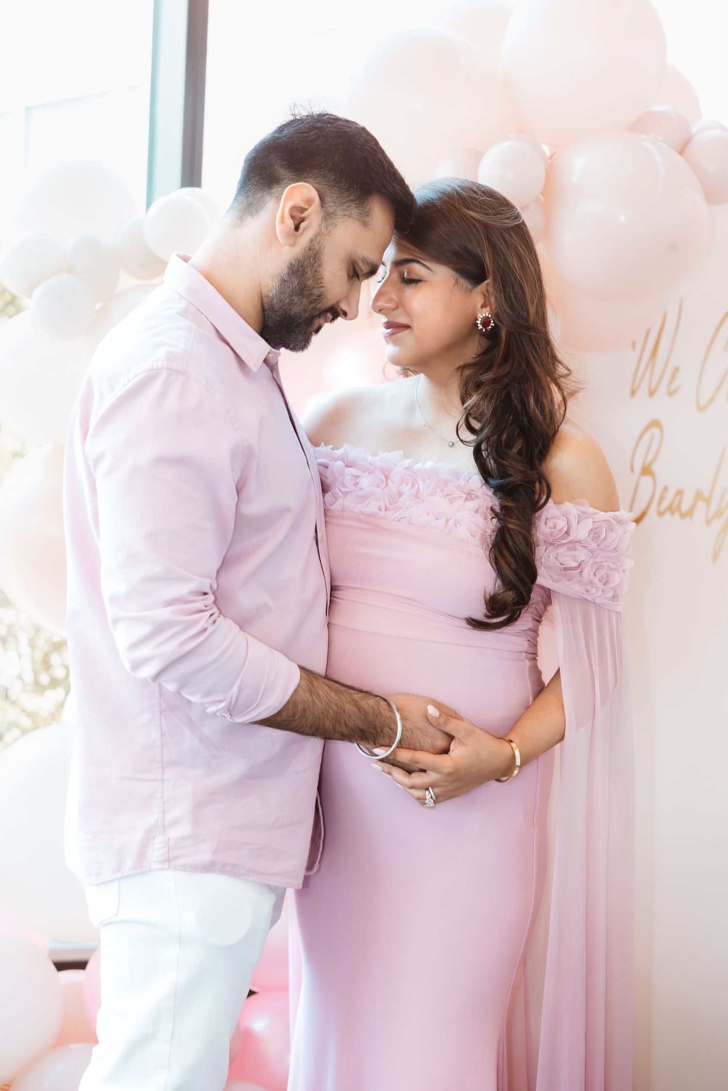 Expecting South Asian couple in matching pink outfits share tender moment at baby shower balloon backdrop.