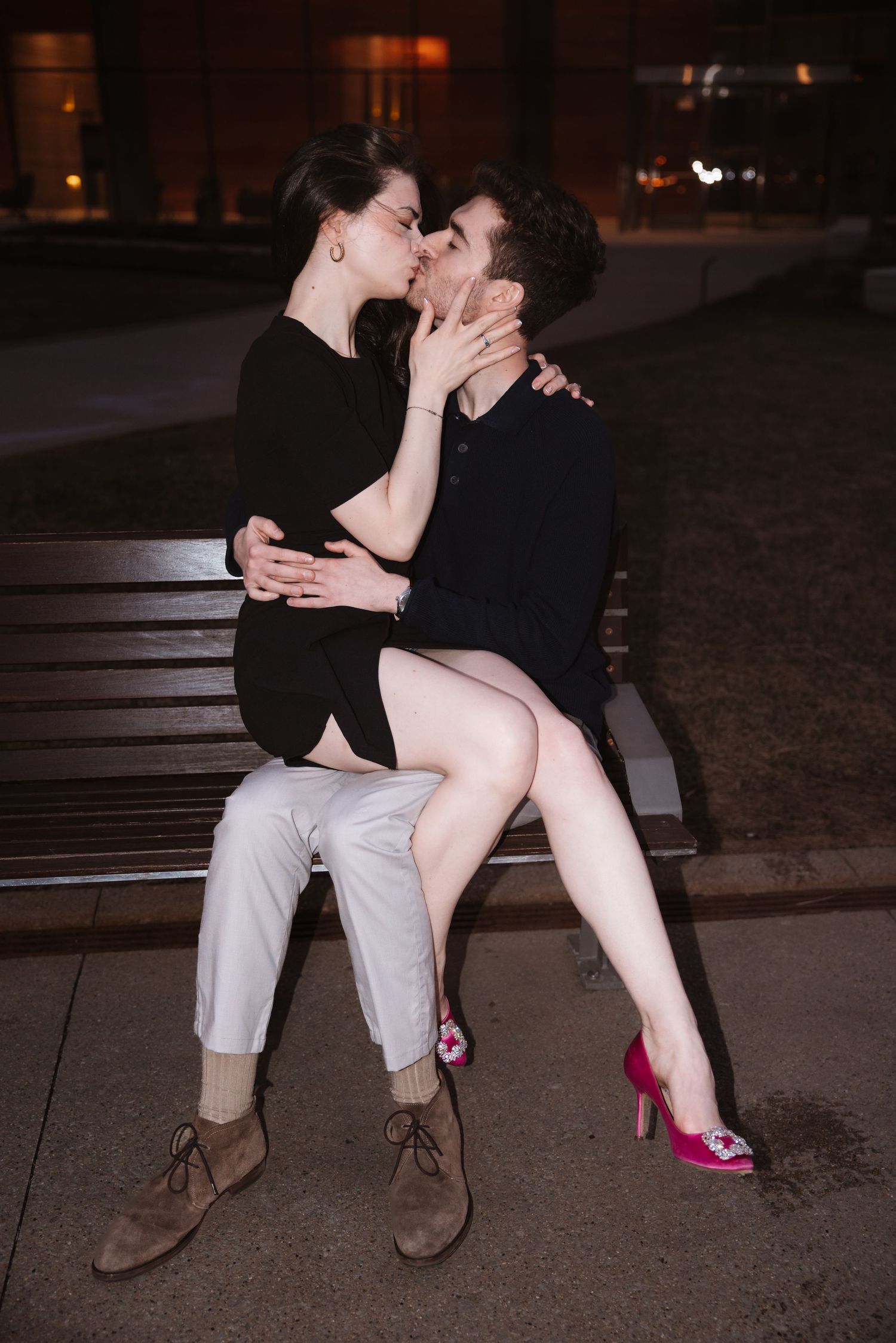 Couple kissing on a bench at night with city lights softly glowing in the background.