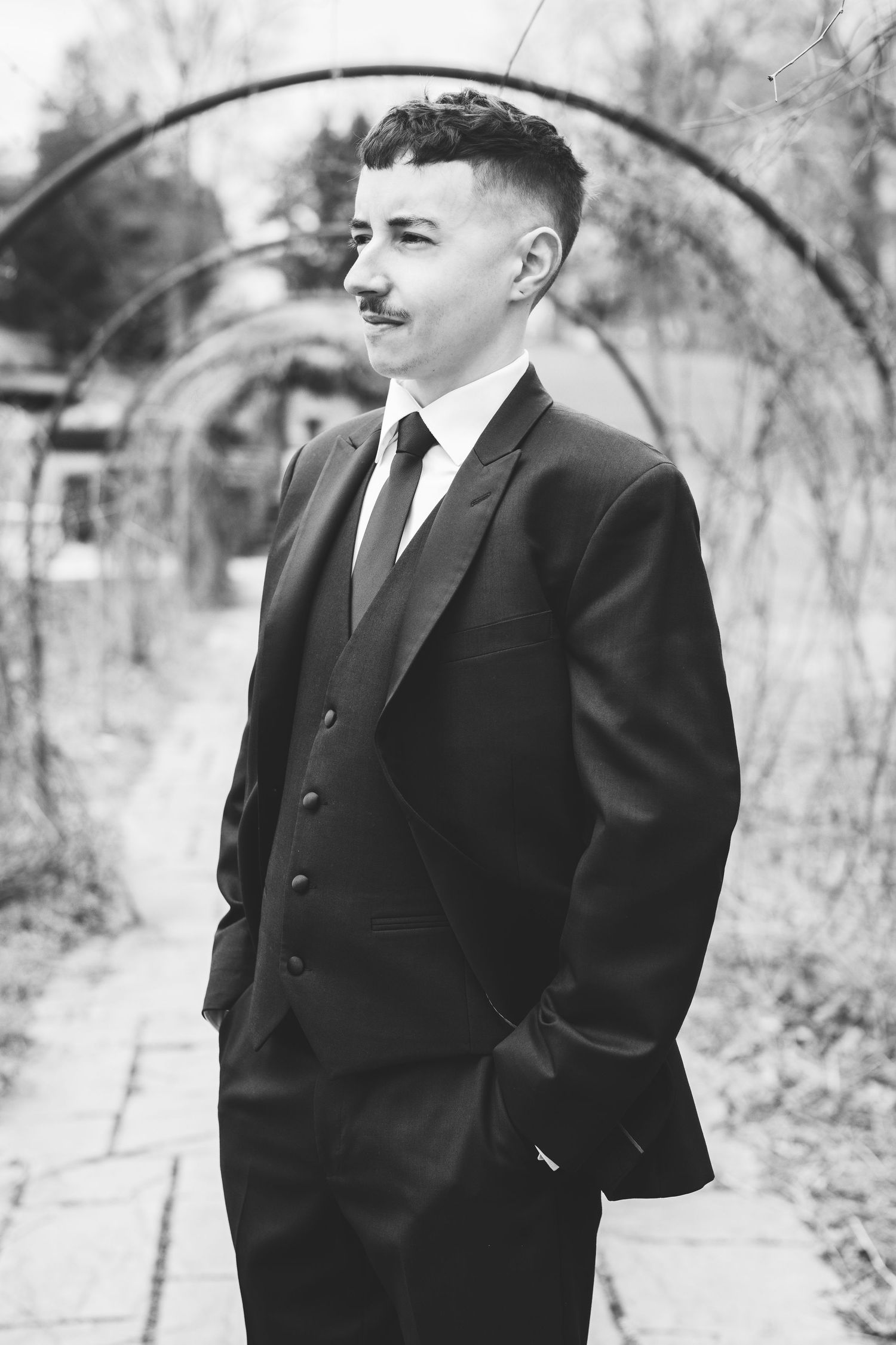 Black and white portrait of a young man in a three-piece suit with tie, standing outdoors near an arched garden trellis.