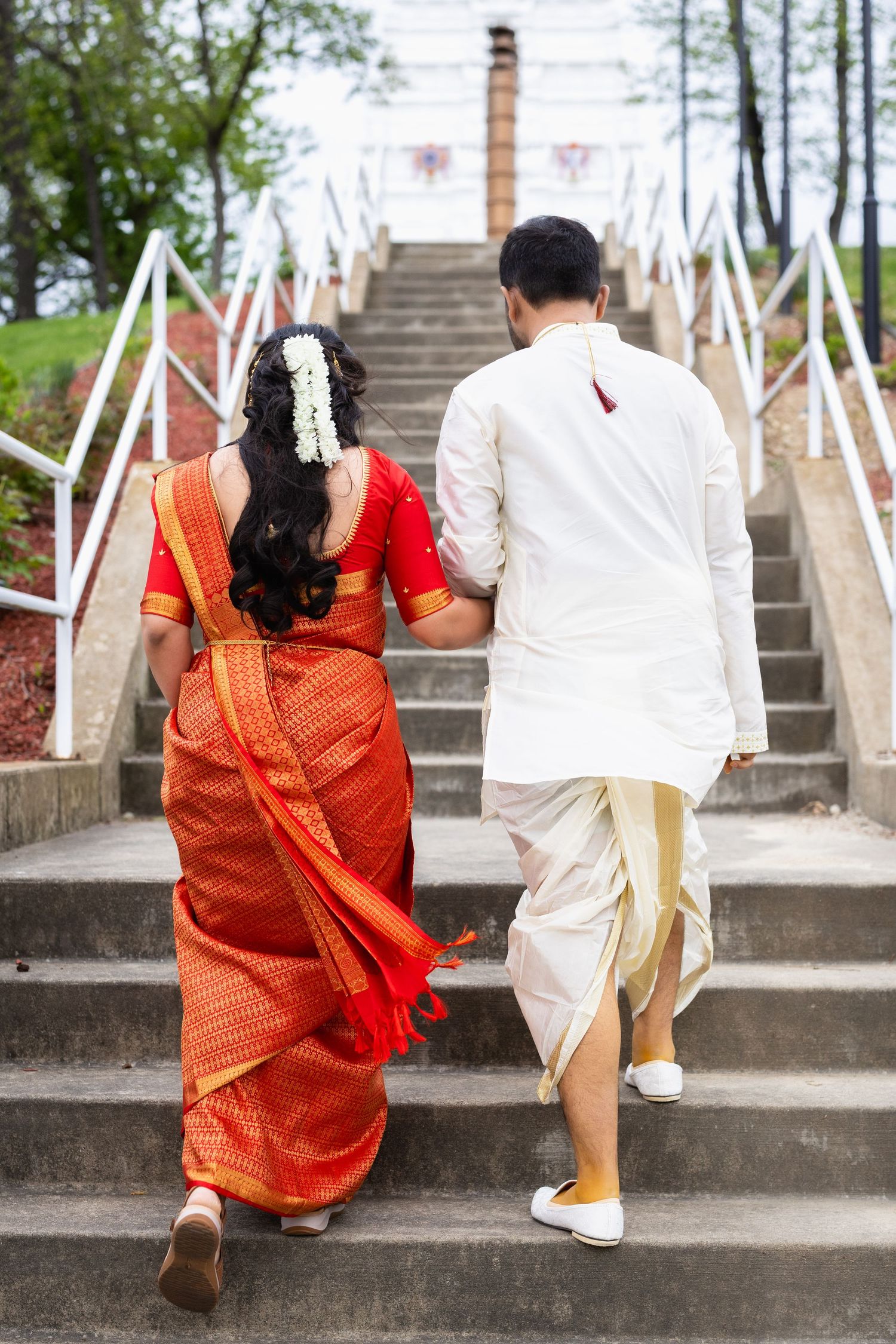 Indian couple in traditional attire walking up temple steps, bride in red saree and groom in white kurta.
