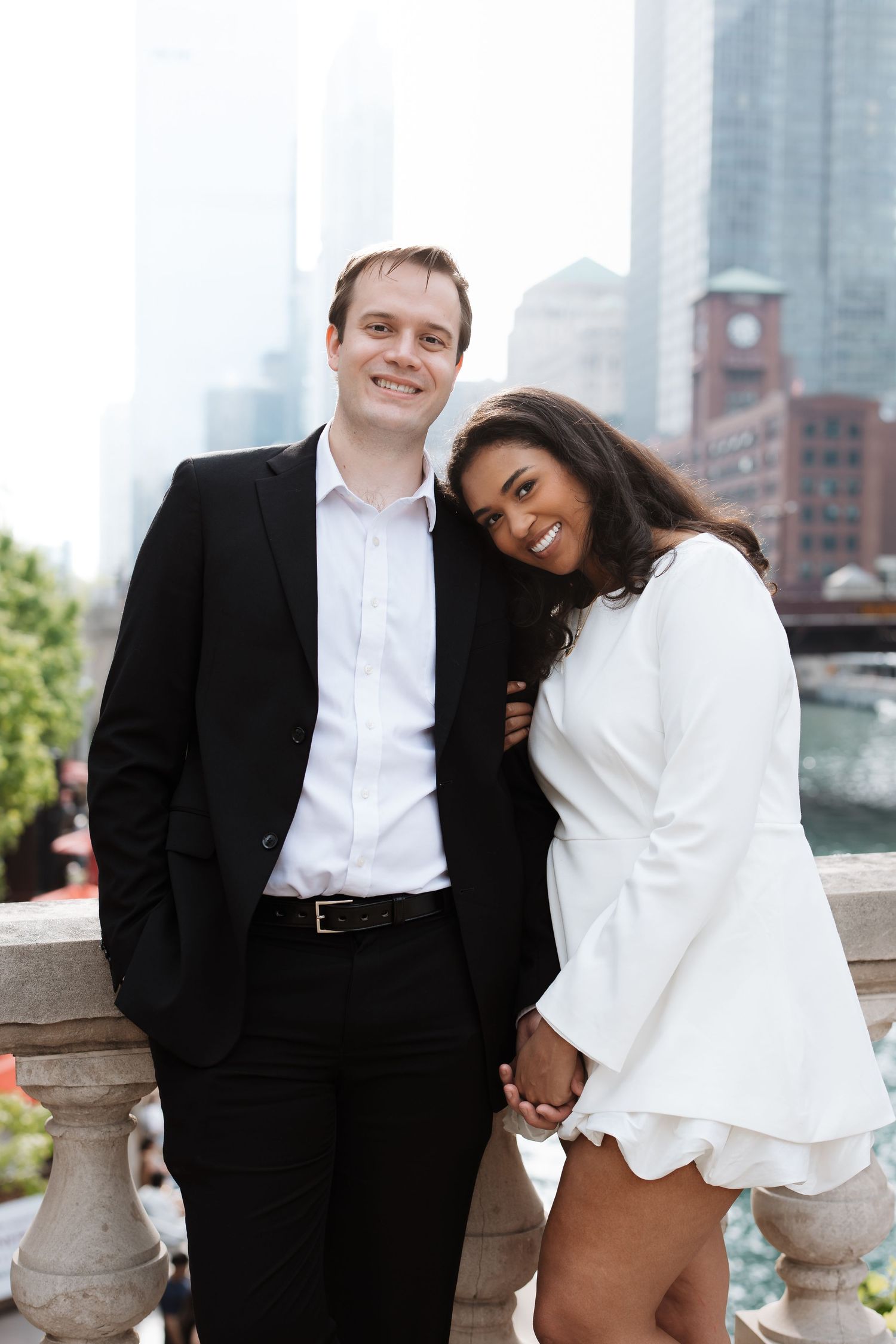 Stylish couple posing hand-in-hand near a stone railing with Chicago skyscrapers in the background.