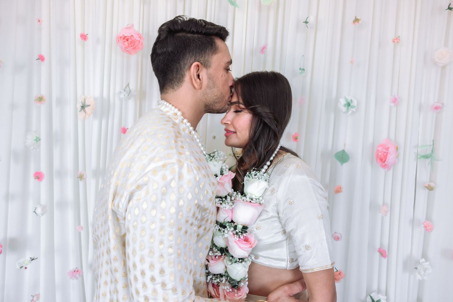 Couple sharing an intimate moment at an Indian engagement ceremony with floral garlands and pink paper flower backdrop.