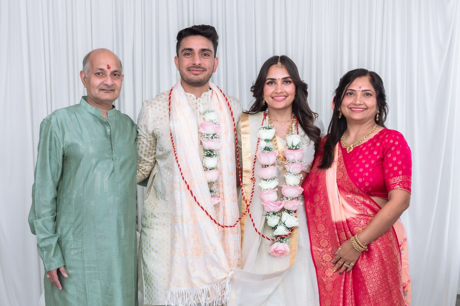 Indian wedding couple wearing floral garlands stands with parents in traditional pink saree and sage green kurta attire.