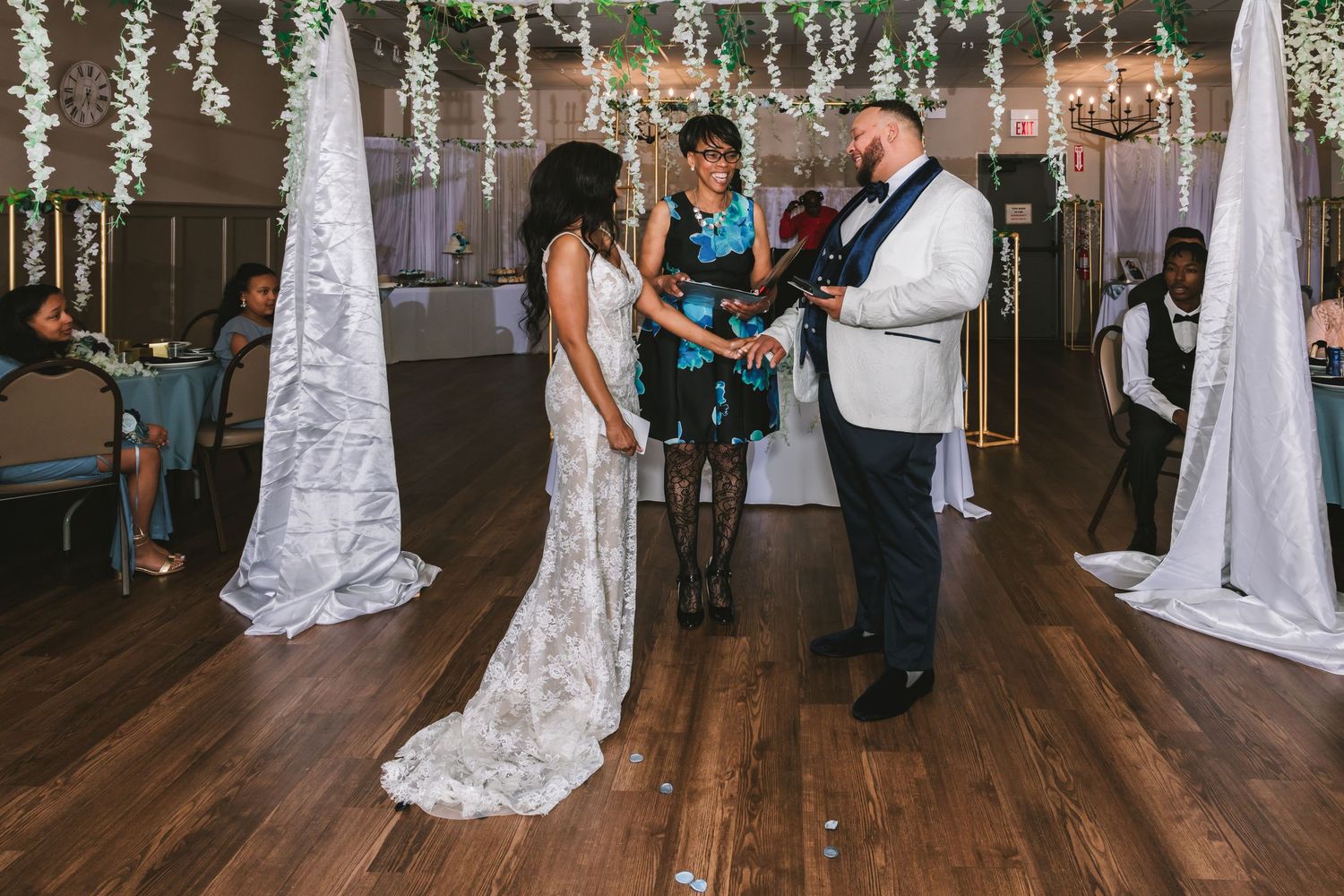 Bride and groom exchange vows with officiant at elegant indoor wedding ceremony with white draping and greenery.
