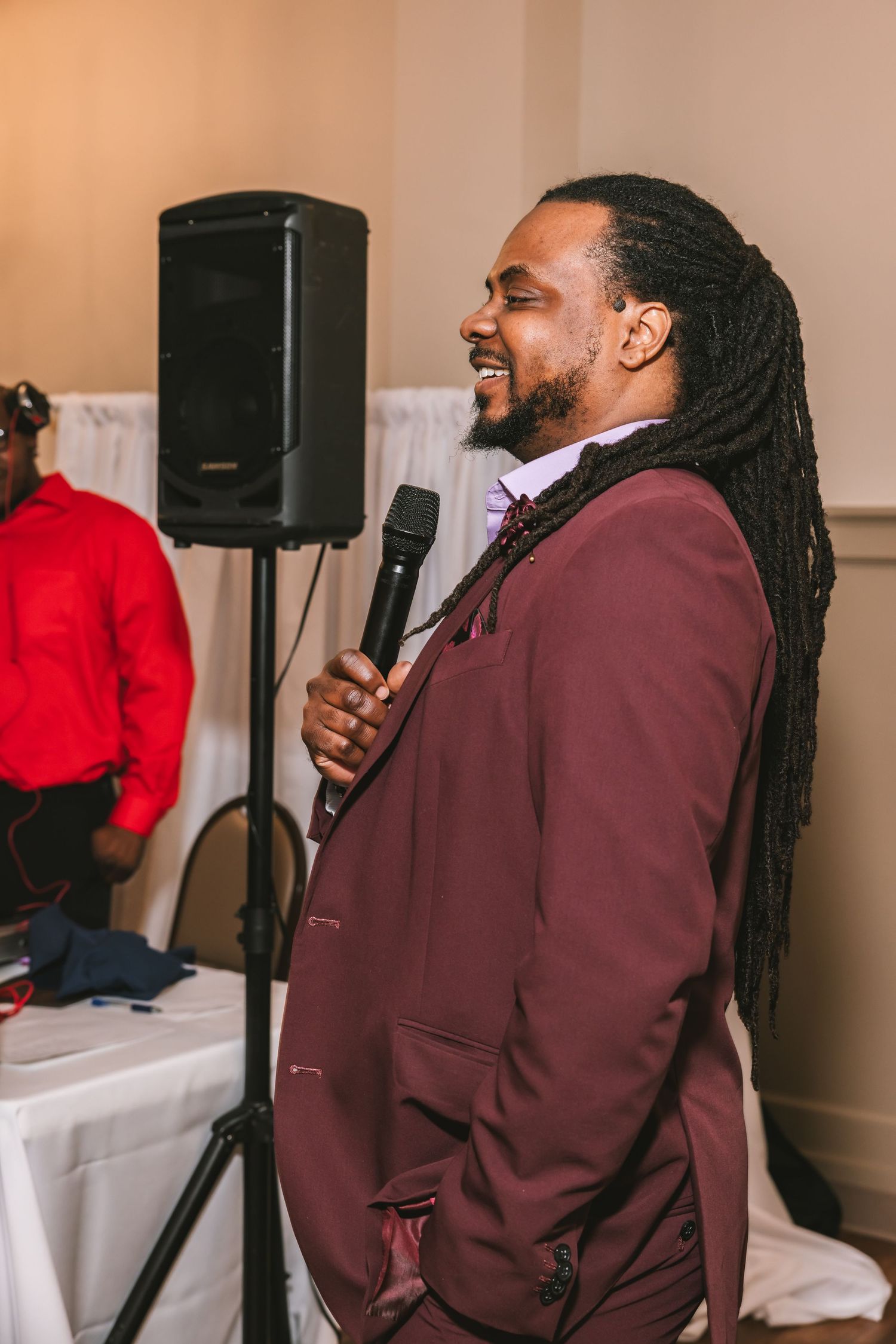 A man in a burgundy suit with long dreadlocks speaks into a microphone at a wedding event.