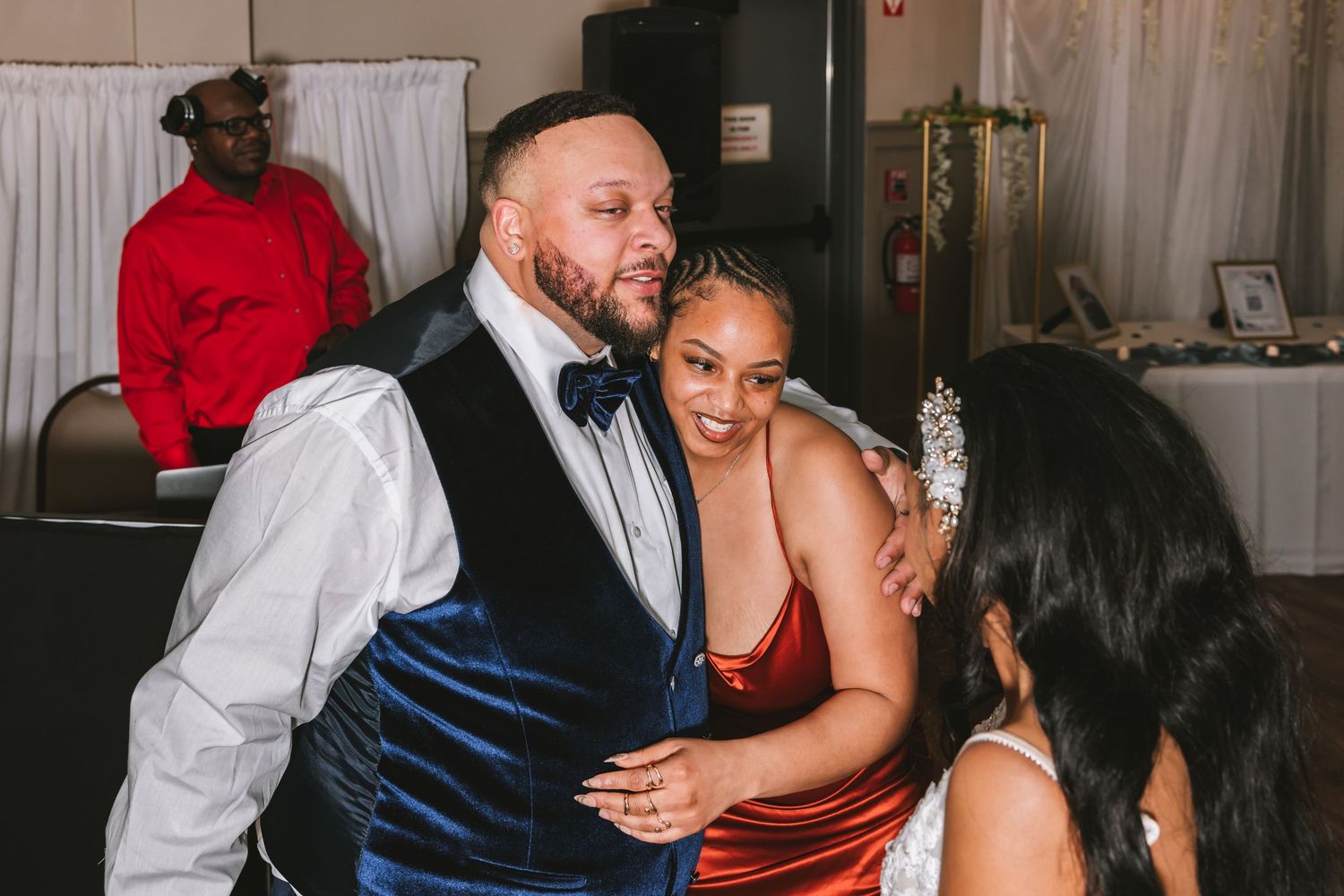 Groom in blue velvet vest embracing smiling guest in red dress at wedding reception.