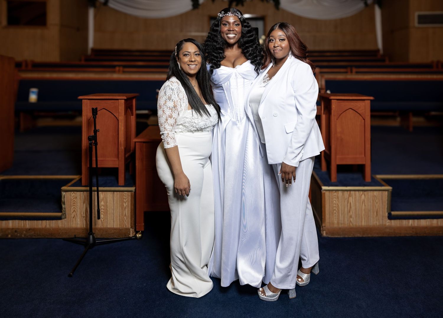 Bride in flowing white gown posing with two women in white outfits inside a decorated church.
