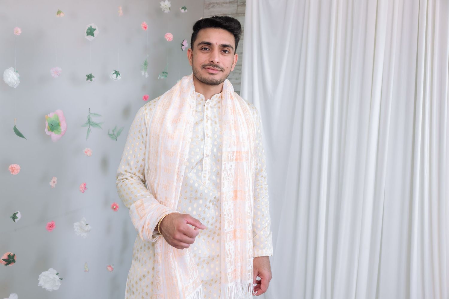 Groom in white and pink sherwani posing confidently before Indian wedding ceremony with floral backdrop.