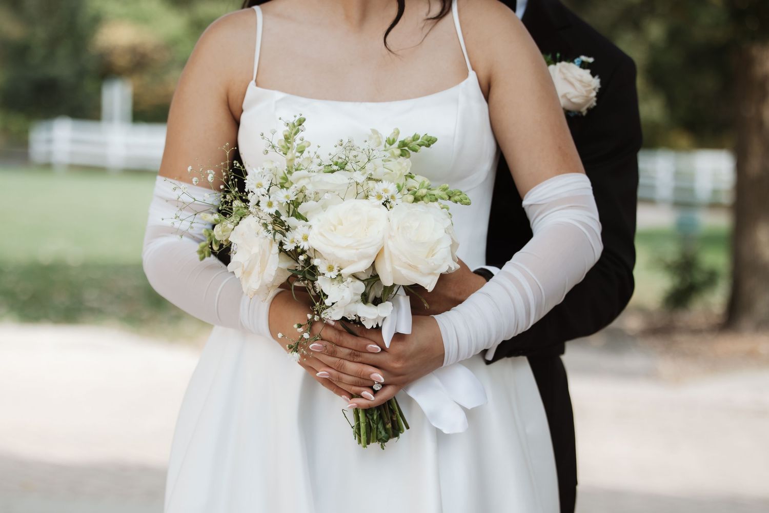 Close-up of bride holding white rose bouquet with groom embracing her from behind outdoors.