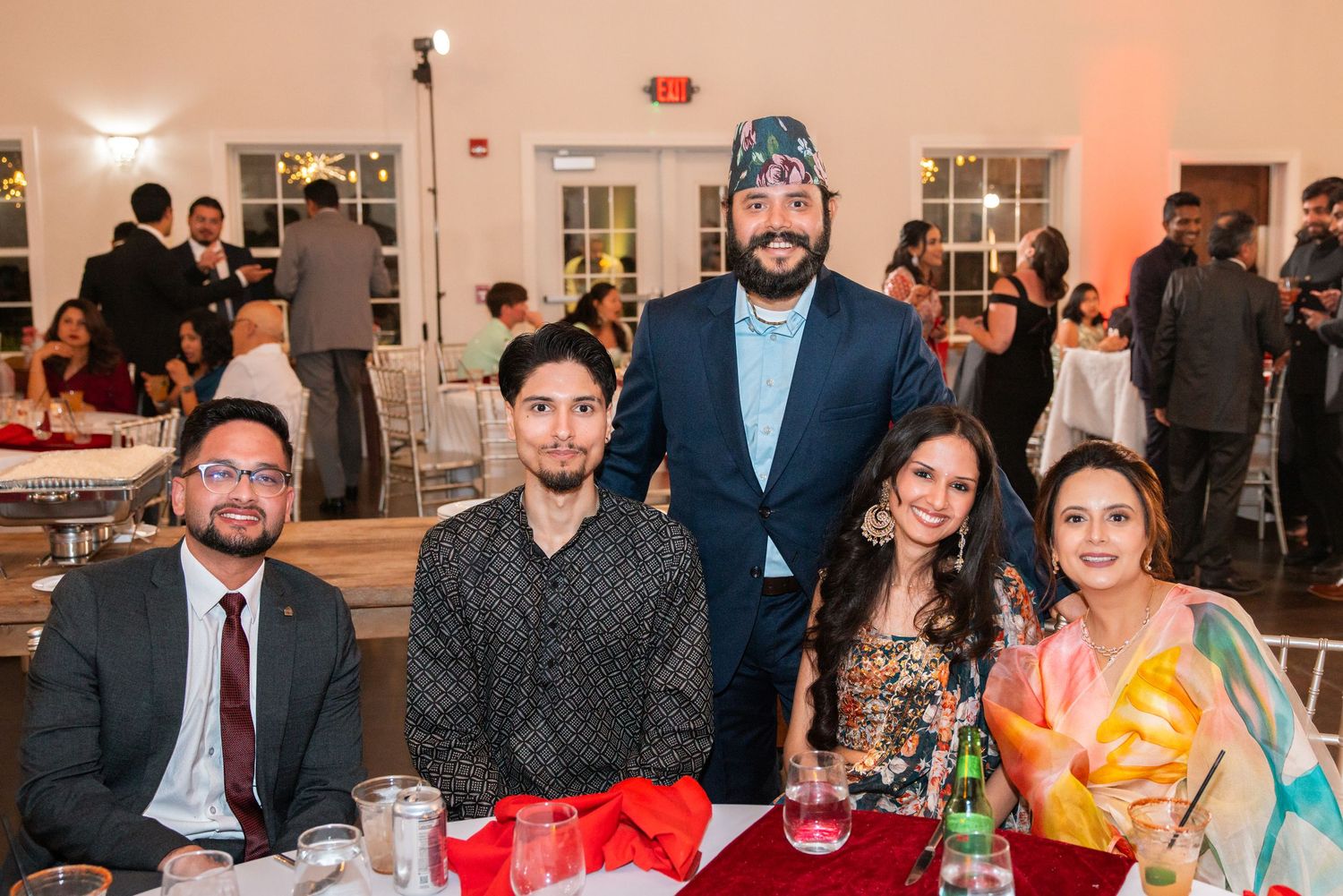 Group of five smiling guests at a South Asian wedding reception seated at a festive dinner table.