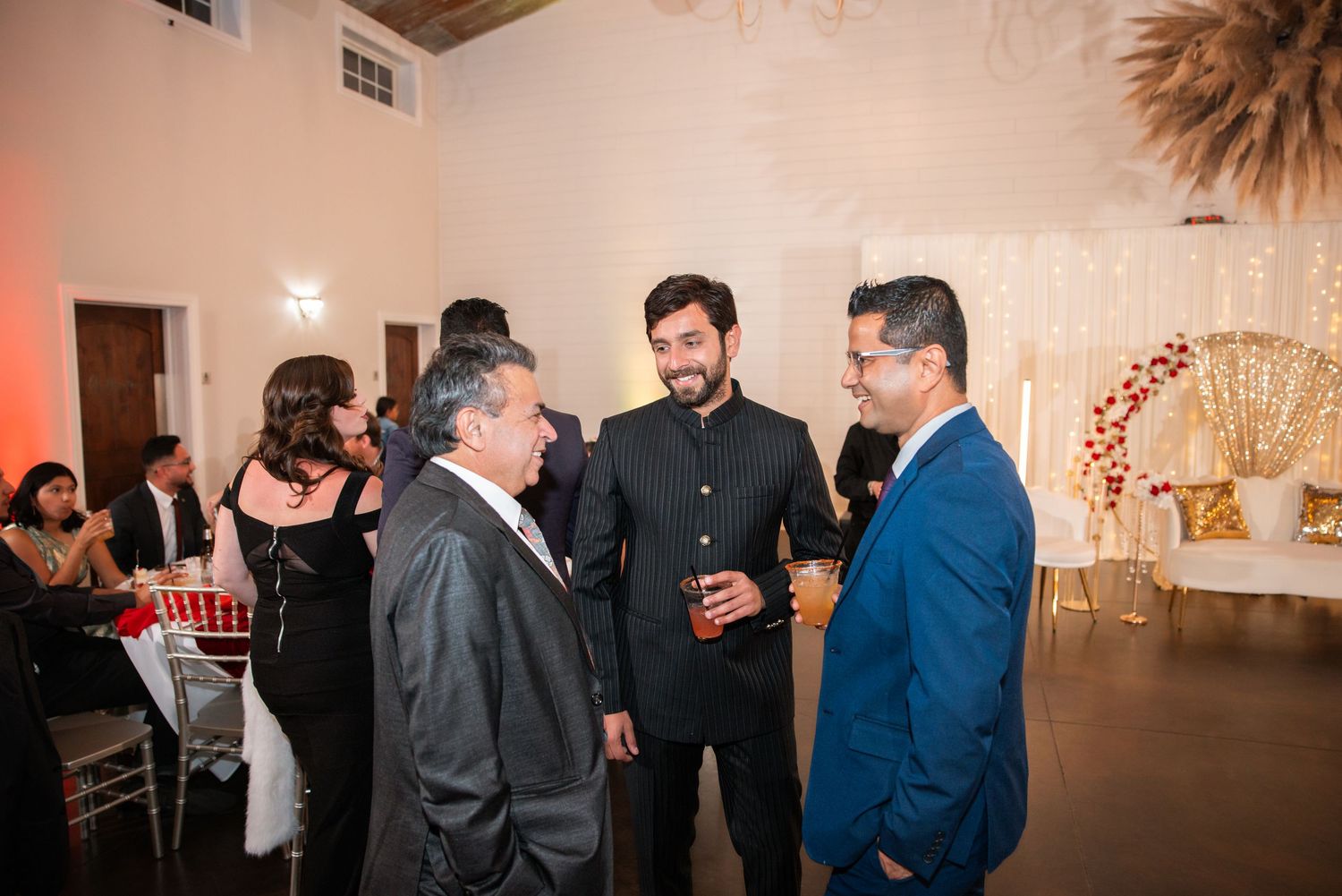 Three men in formal attire engaged in conversation at an elegant Indian wedding reception venue.