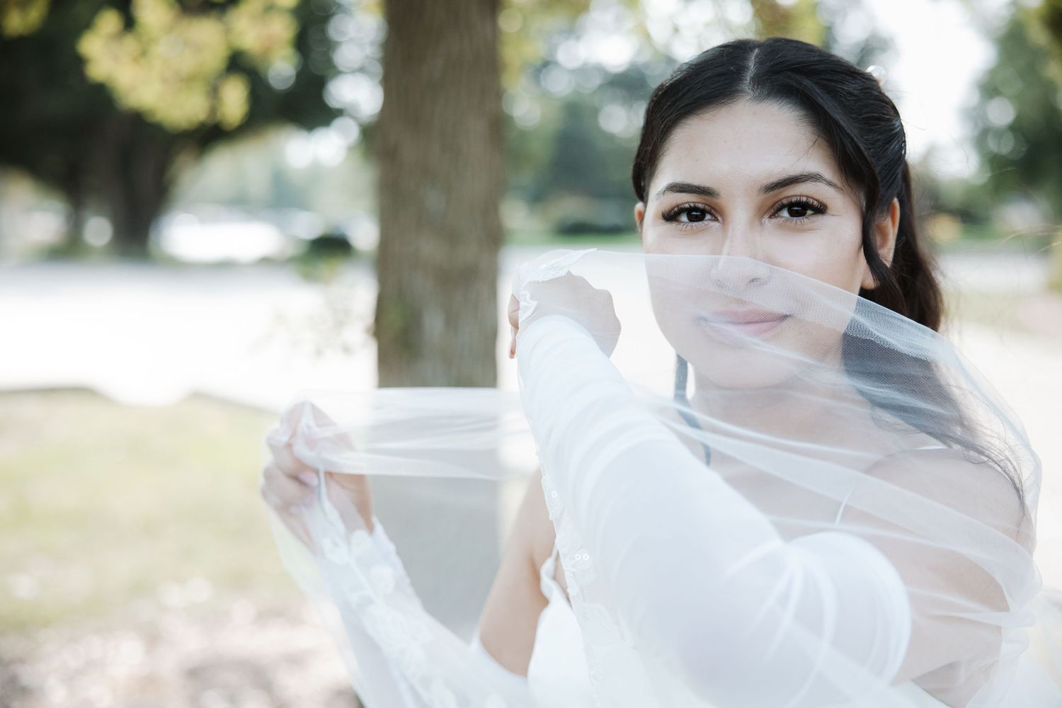 Bride in white long-sleeve gown holding flowing veil outdoors under sunlit trees.