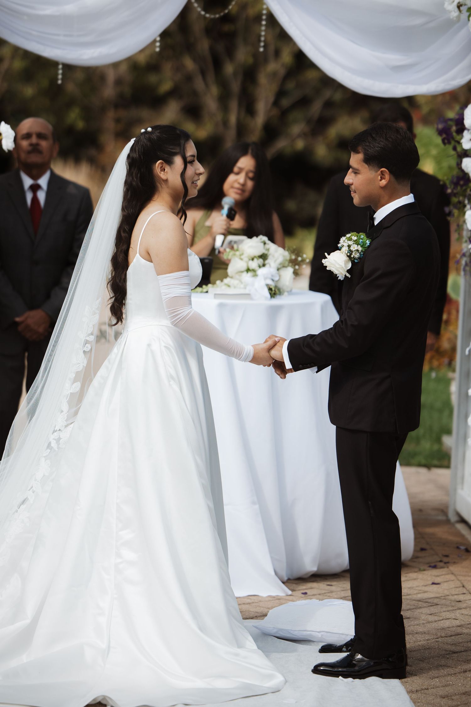 Bride in white gown and groom in black tux hold hands at outdoor wedding altar.