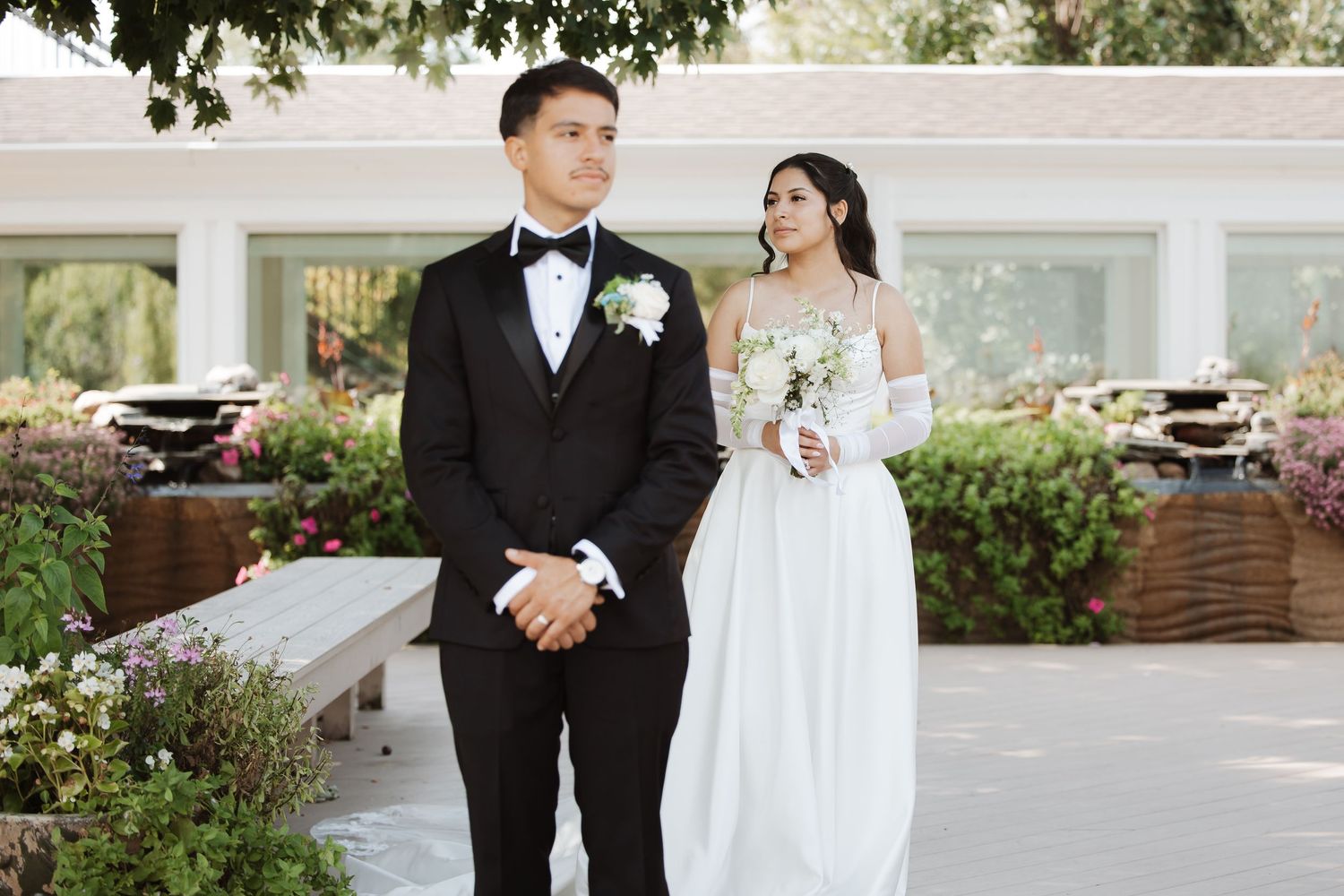 Wedding couple in formal attire stand together outdoors, bride in white gown with gloves holding white bouquet.