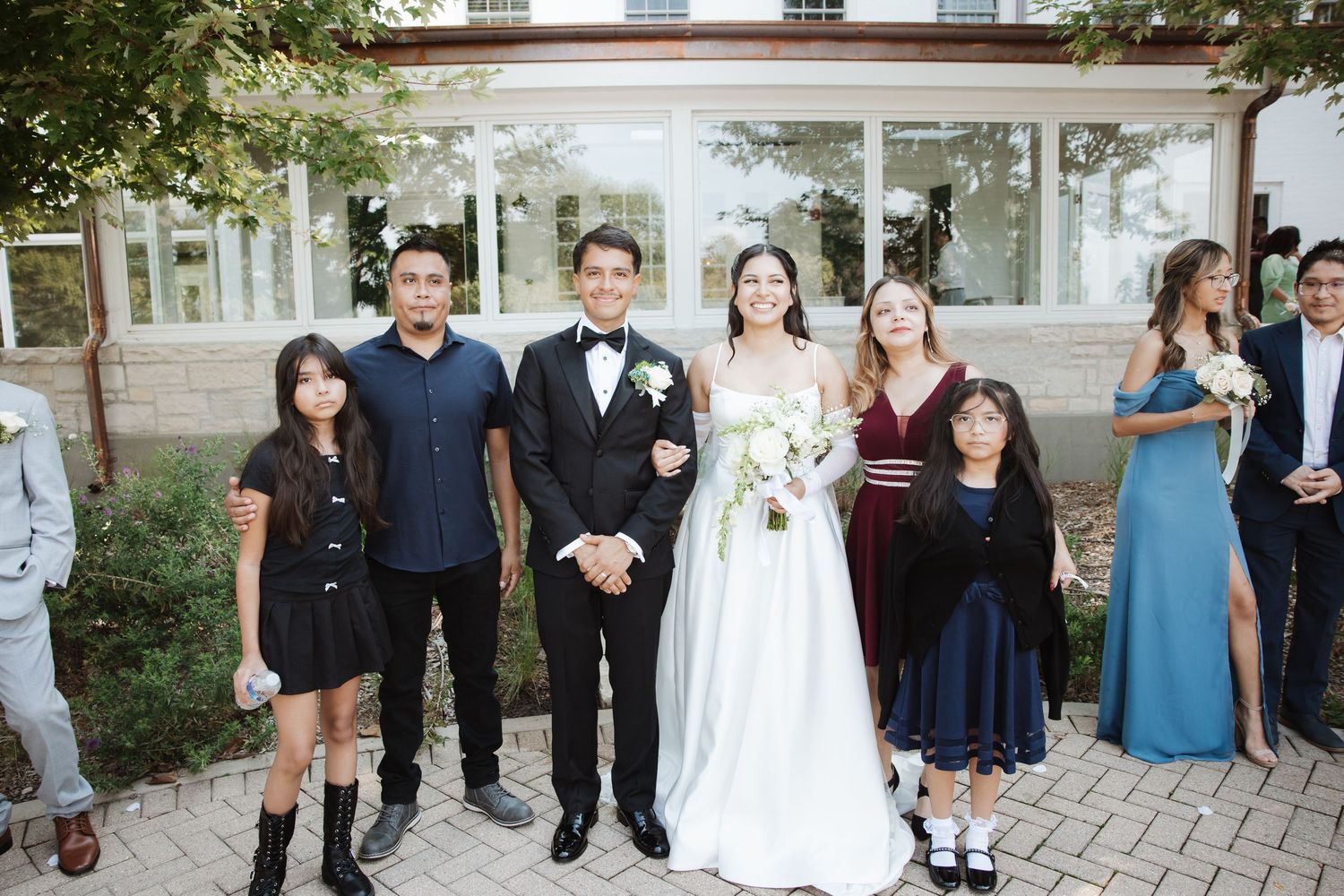 Bride and groom stand with four family members including young girl in navy dress outside venue.
