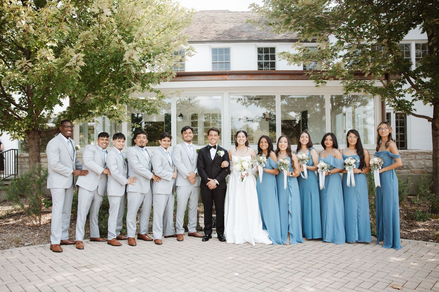 Large wedding party stands outside white venue, groomsmen in light gray suits and bridesmaids in dusty blue gowns.