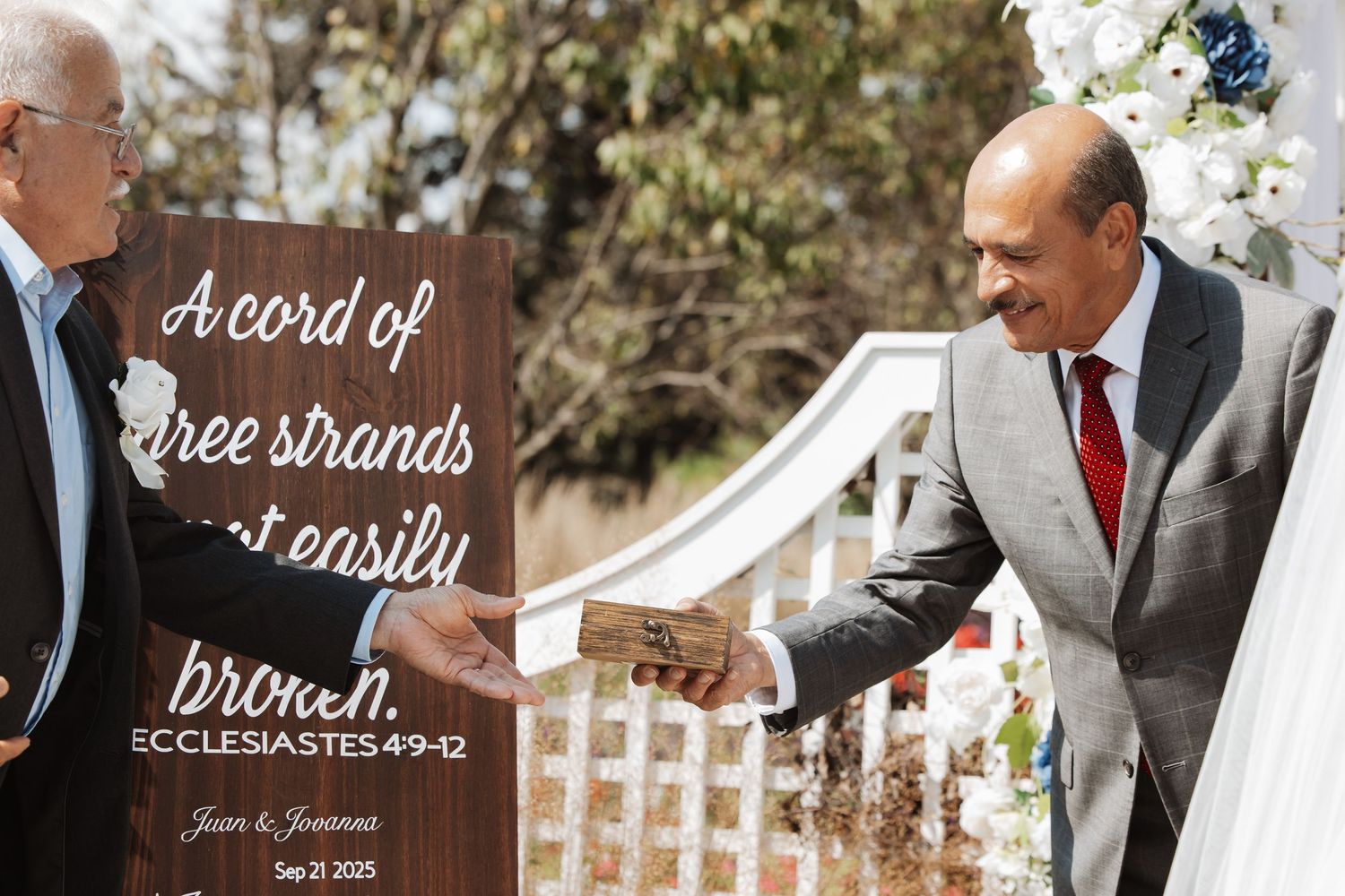 Man presents wooden sign quoting Ecclesiastes 4:9-12 at outdoor wedding ceremony.