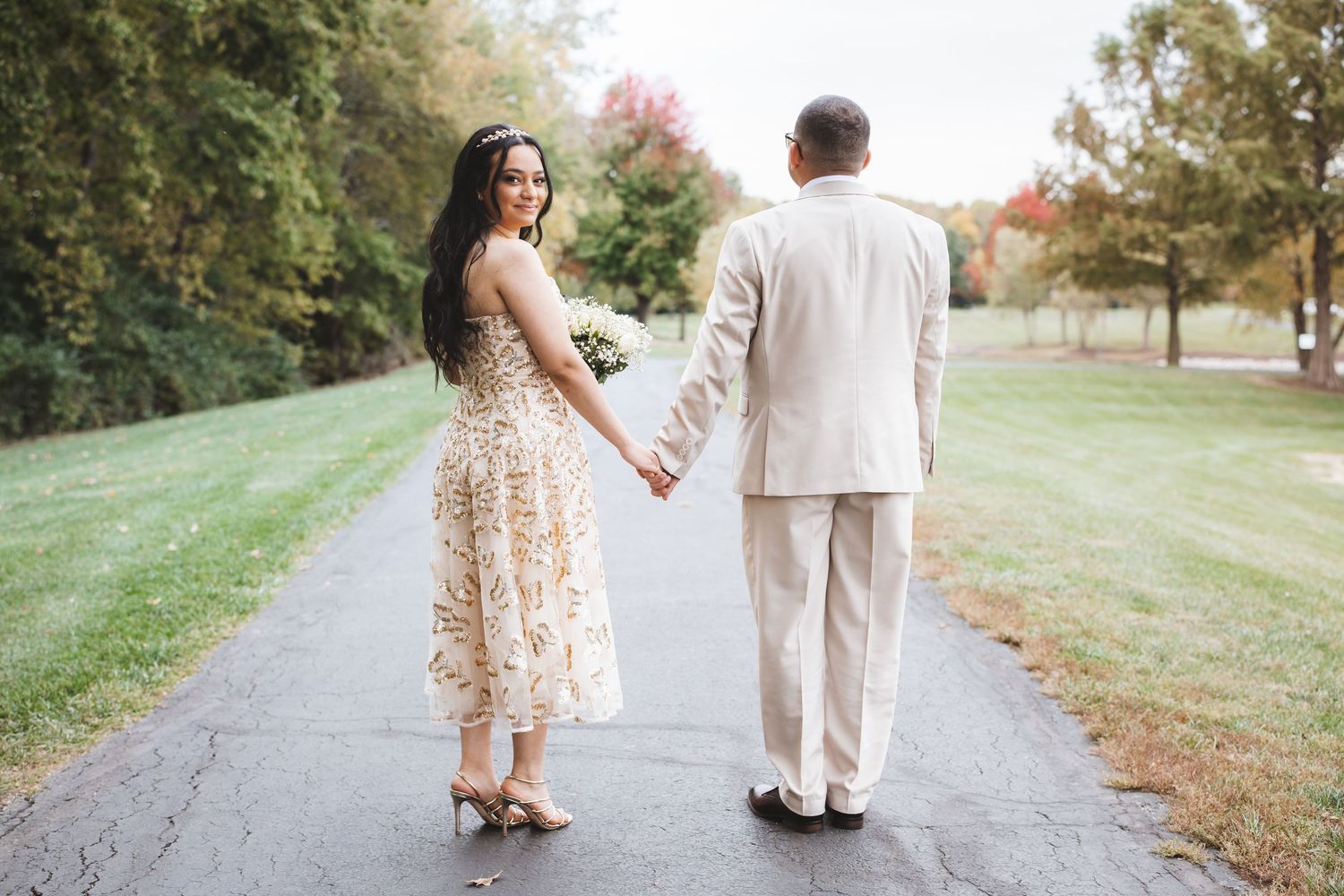 Bride looking back smiling while holding groom's hand walking on a paved path surrounded by autumn trees.