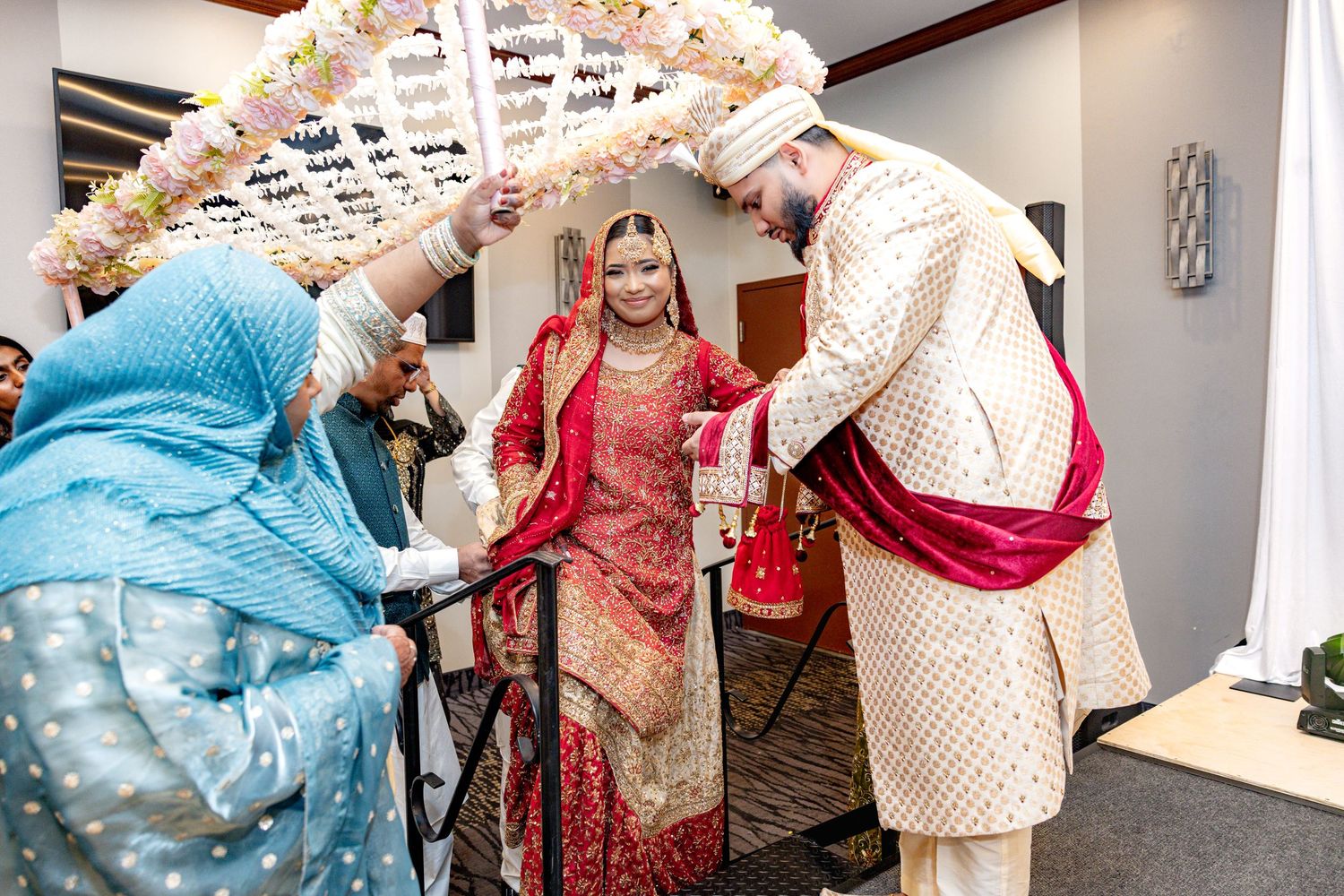 South Asian bride in red lehenga and groom in cream sherwani entering under flower canopy at wedding.