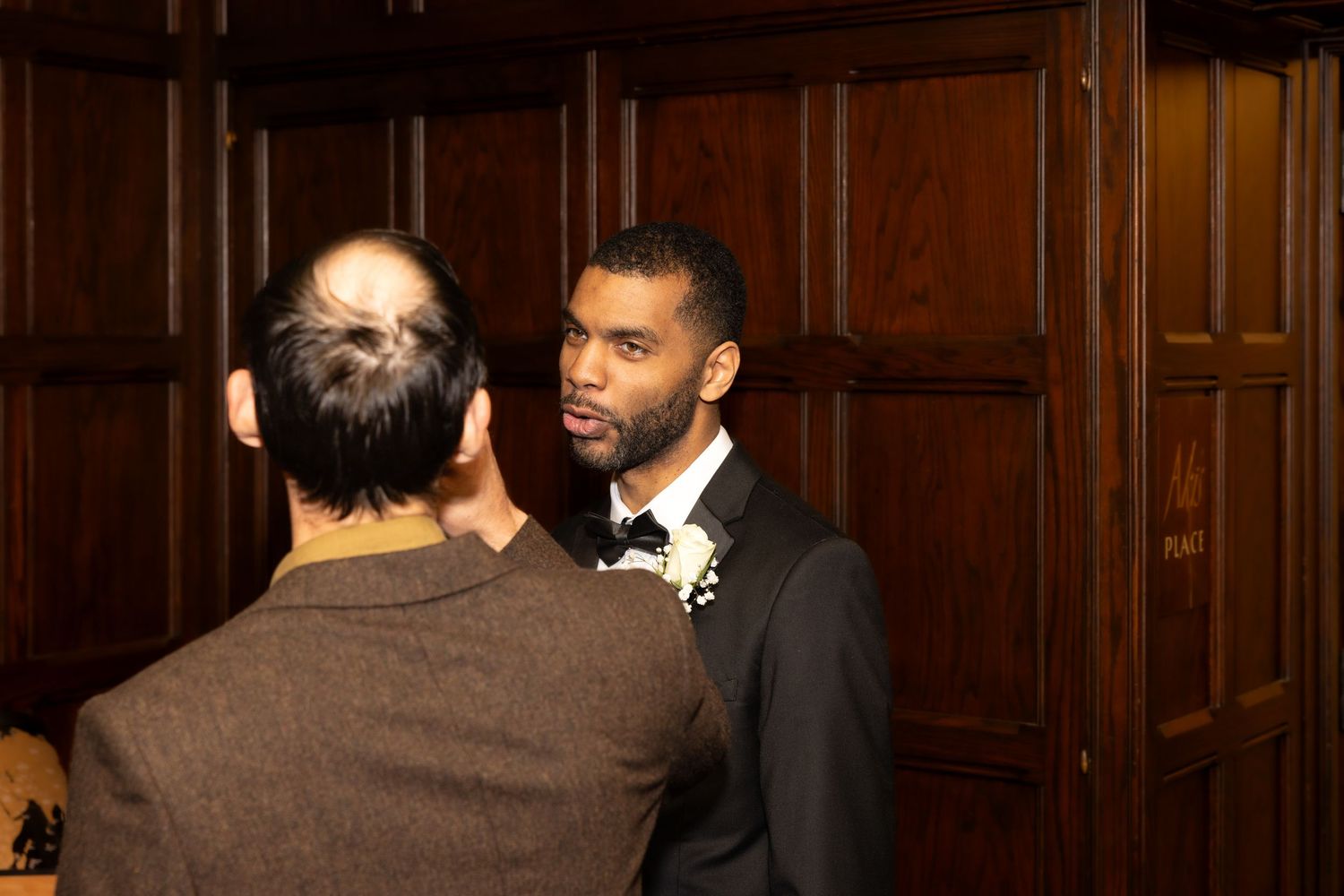 Elegant man in tuxedo with bow tie engaged in conversation at formal wood-paneled venue.