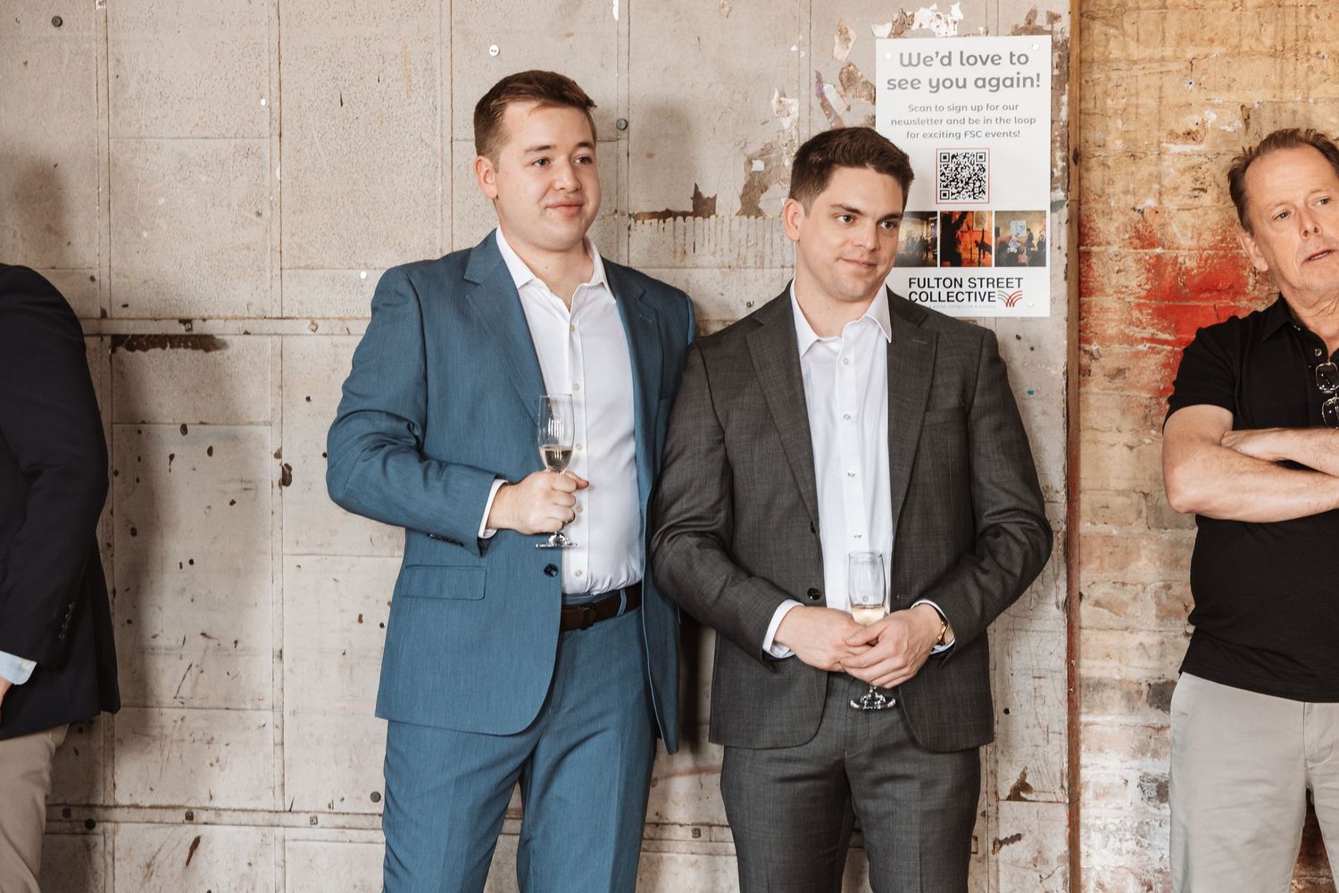 Two men in suits standing against a concrete wall at an indoor social gathering.