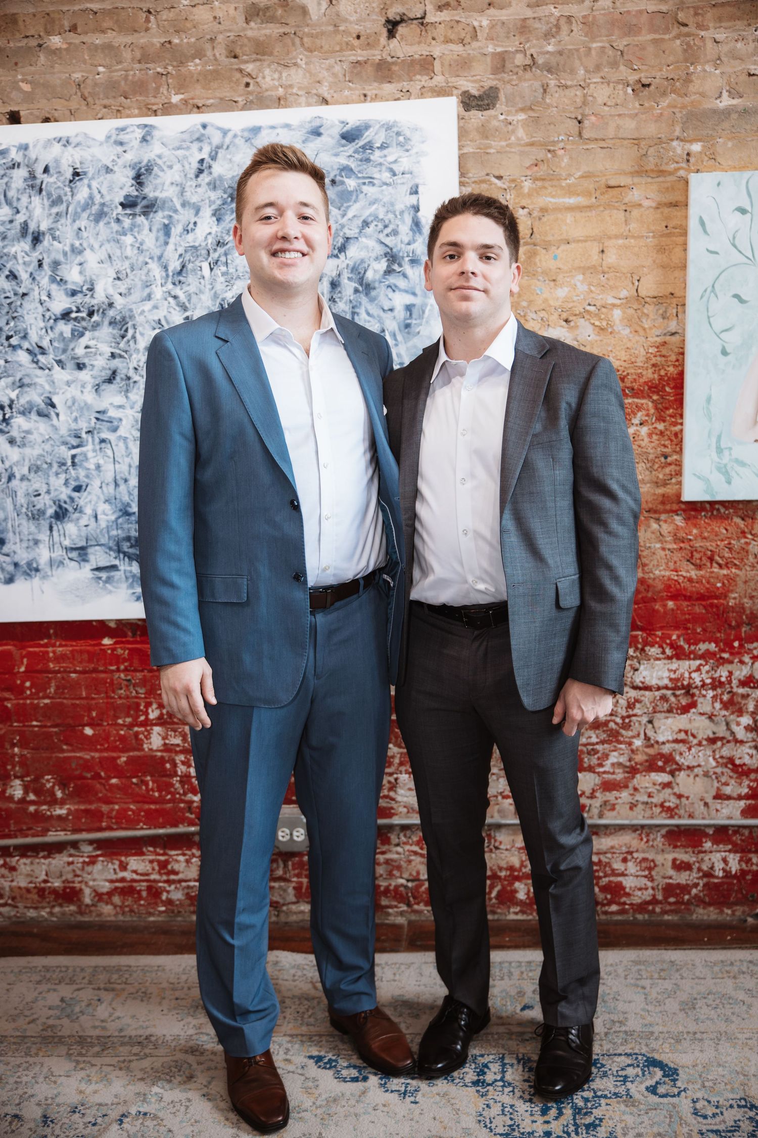 Two men in formal suits smile while posing in an art gallery with exposed brick walls.