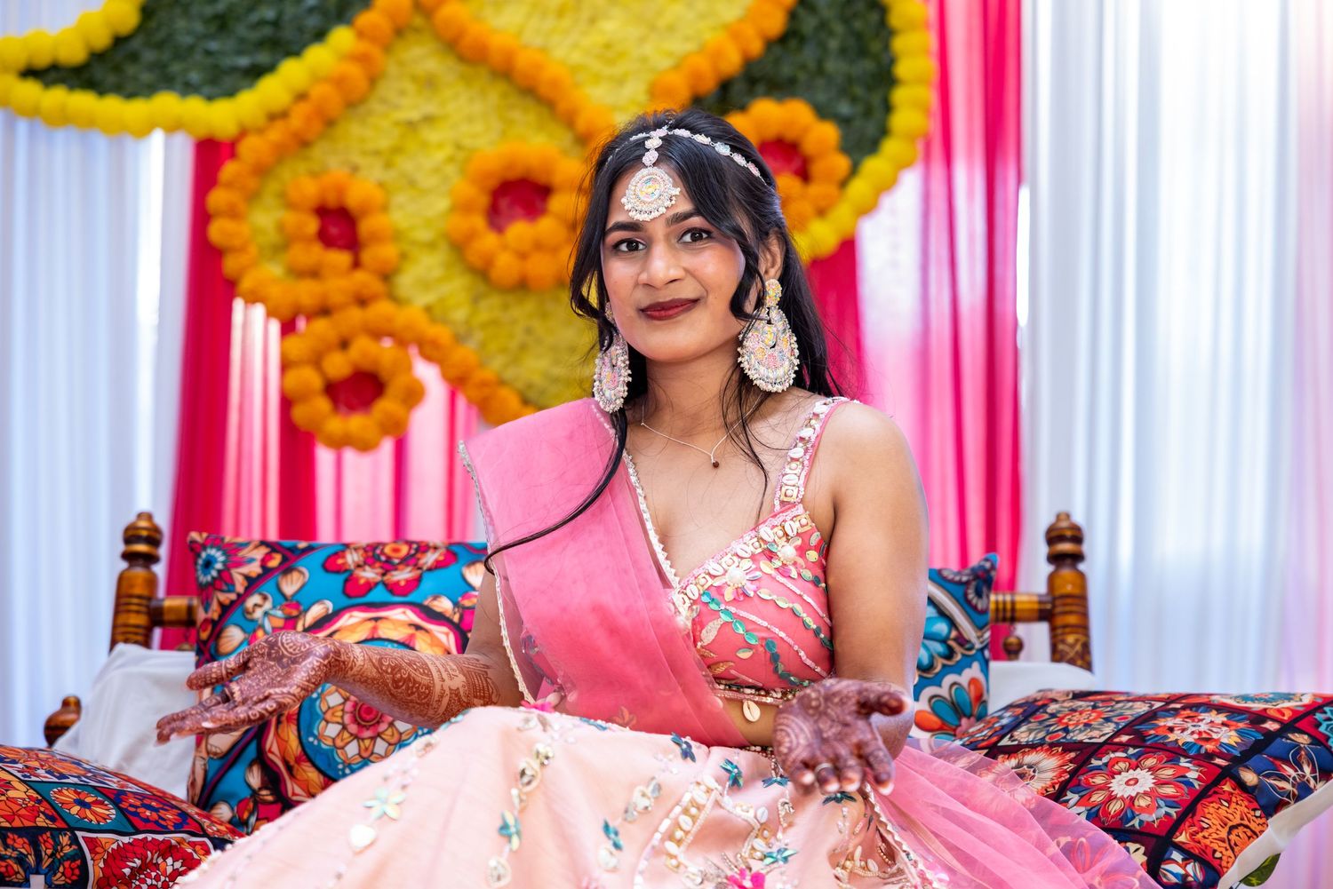 Indian bride in pink lehenga with mehndi hands poses at decorated mehendi ceremony.