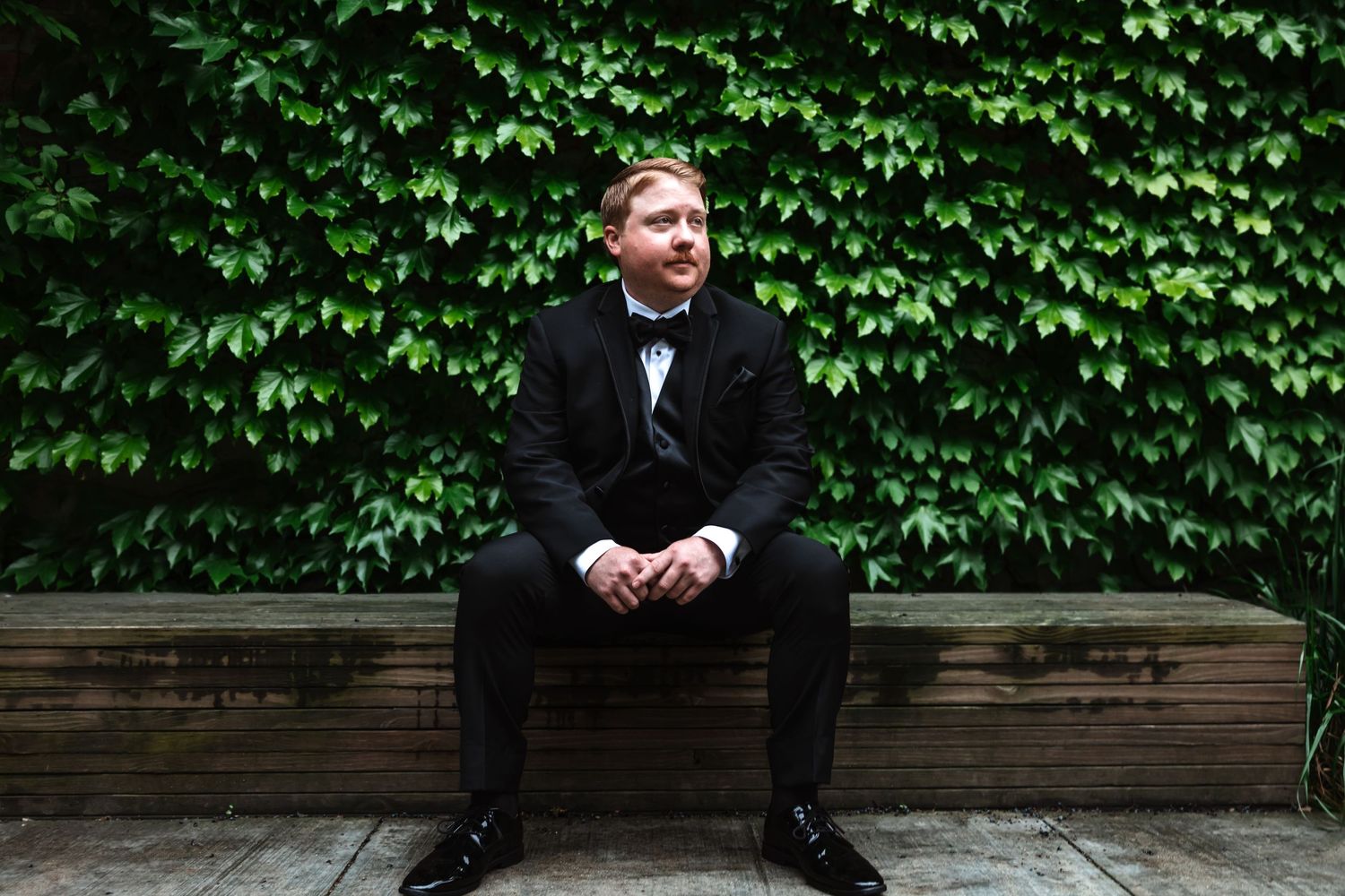 Groom in black tuxedo sitting on wooden bench against lush green ivy wall.