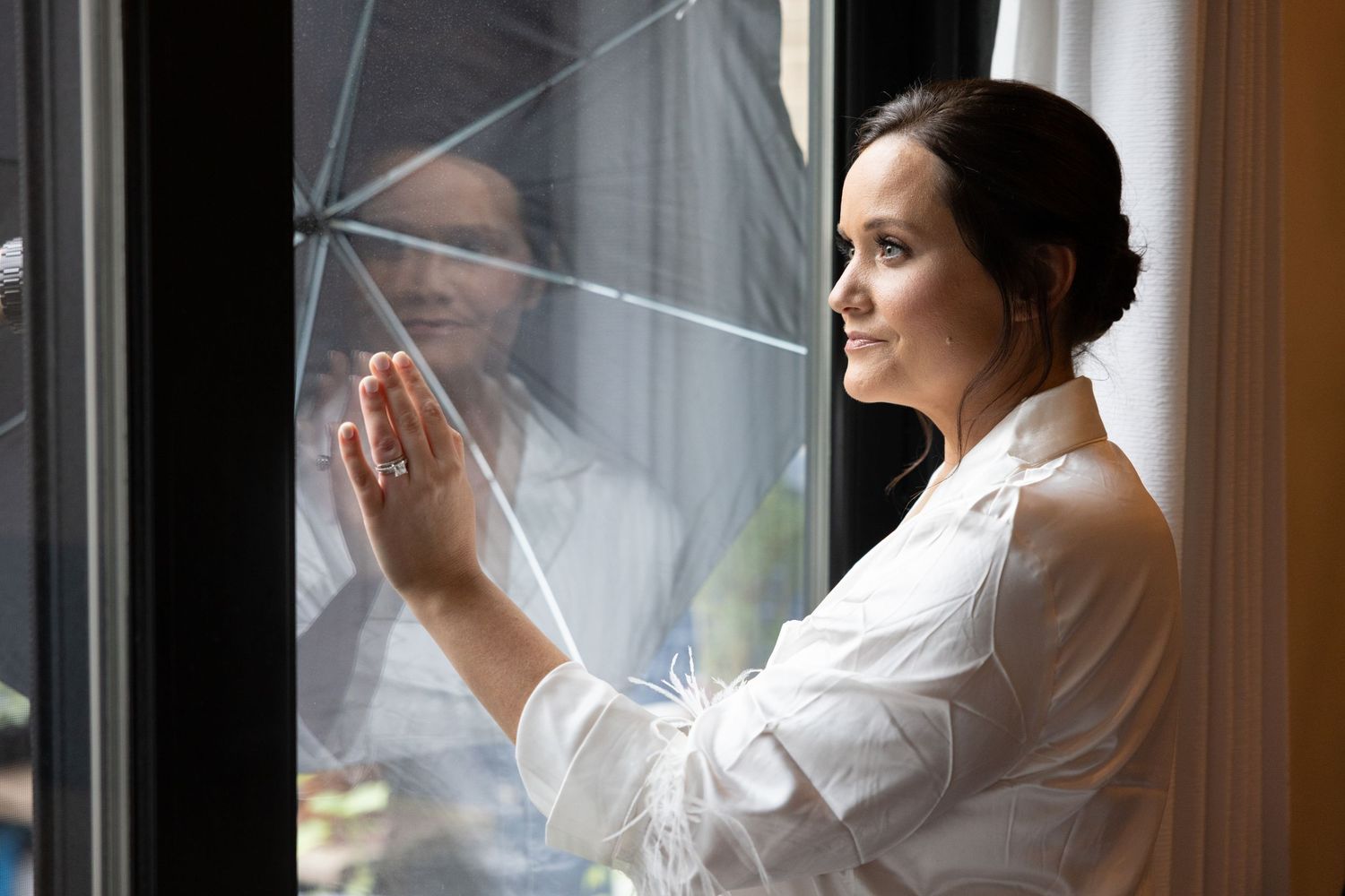 Bride in white robe with diamond ring pressed against sunlit window looking outside.