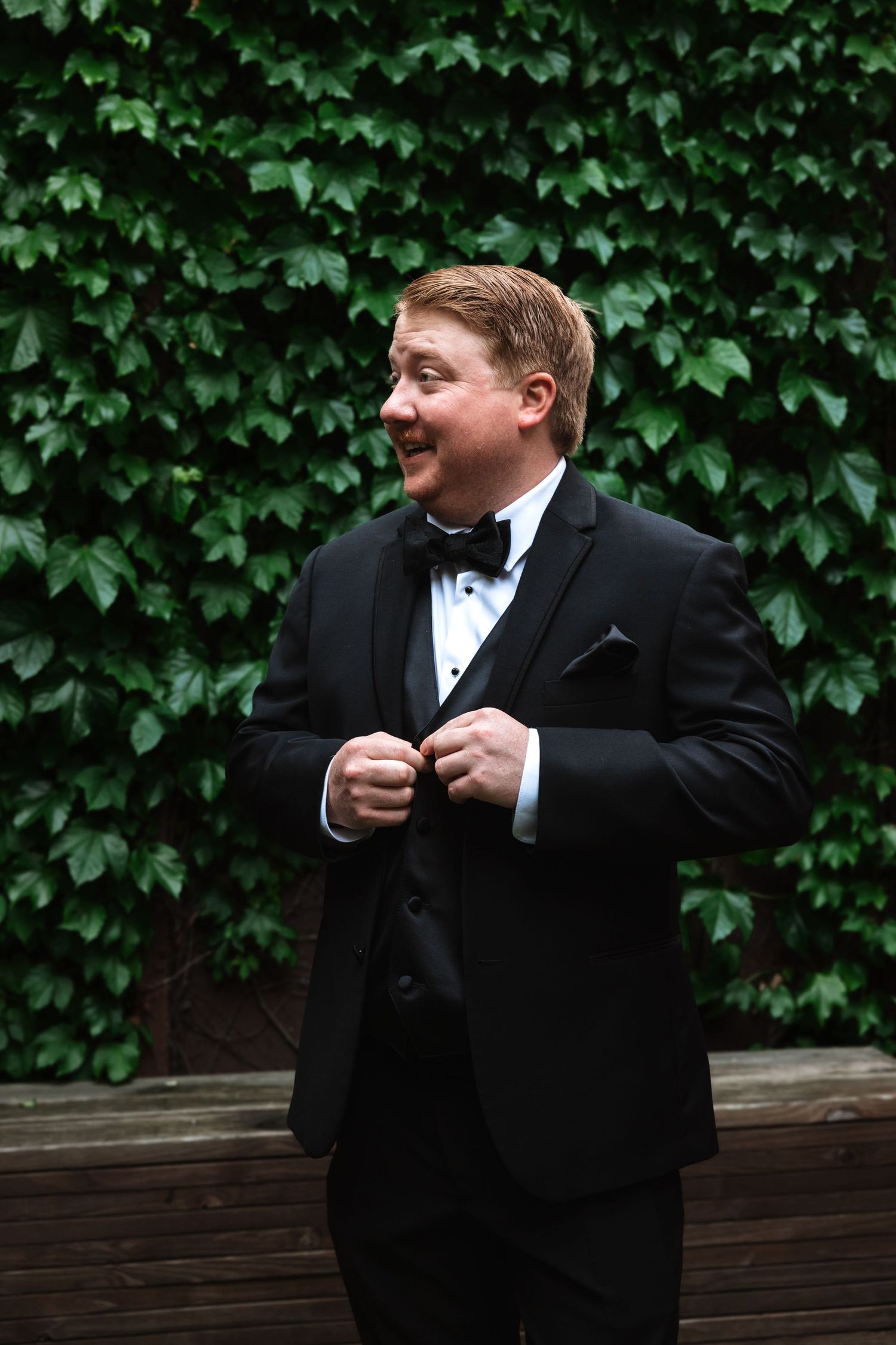 Smiling groom in black tuxedo and bow tie buttoning jacket in front of ivy wall.