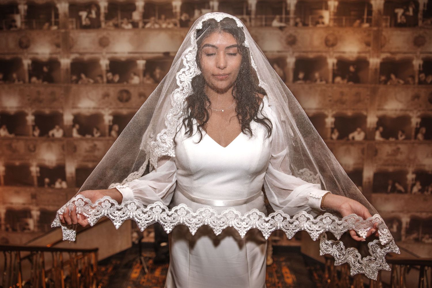 Bride in white satin gown holds lace-trimmed veil in ornate vintage venue.