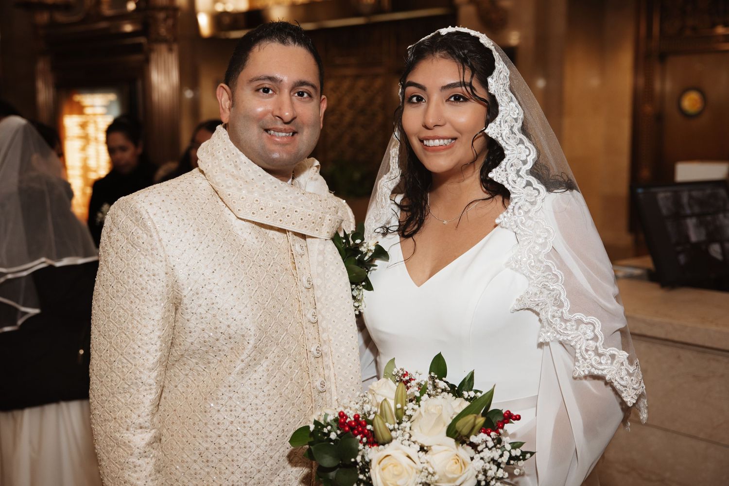 Newlywed couple smiling at camera, groom in embroidered cream sherwani beside bride in white wedding dress.
