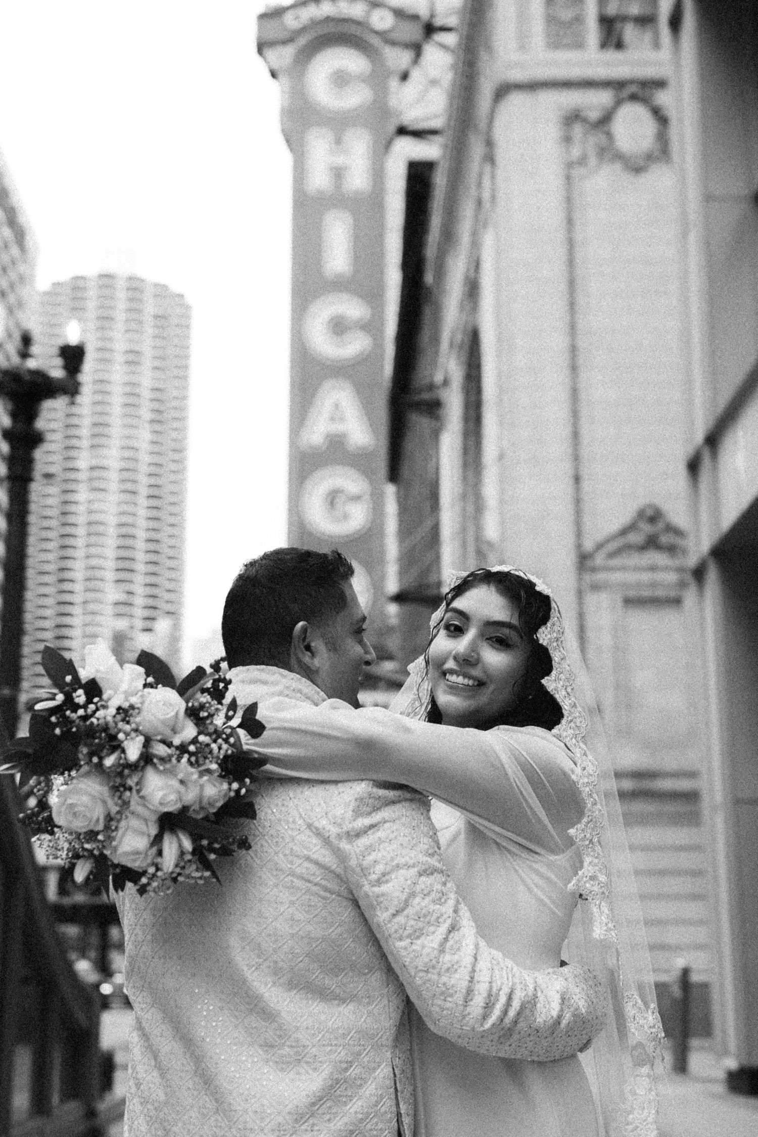 Smiling bride embraces groom holding bouquet beneath Chicago Theatre sign in black and white.