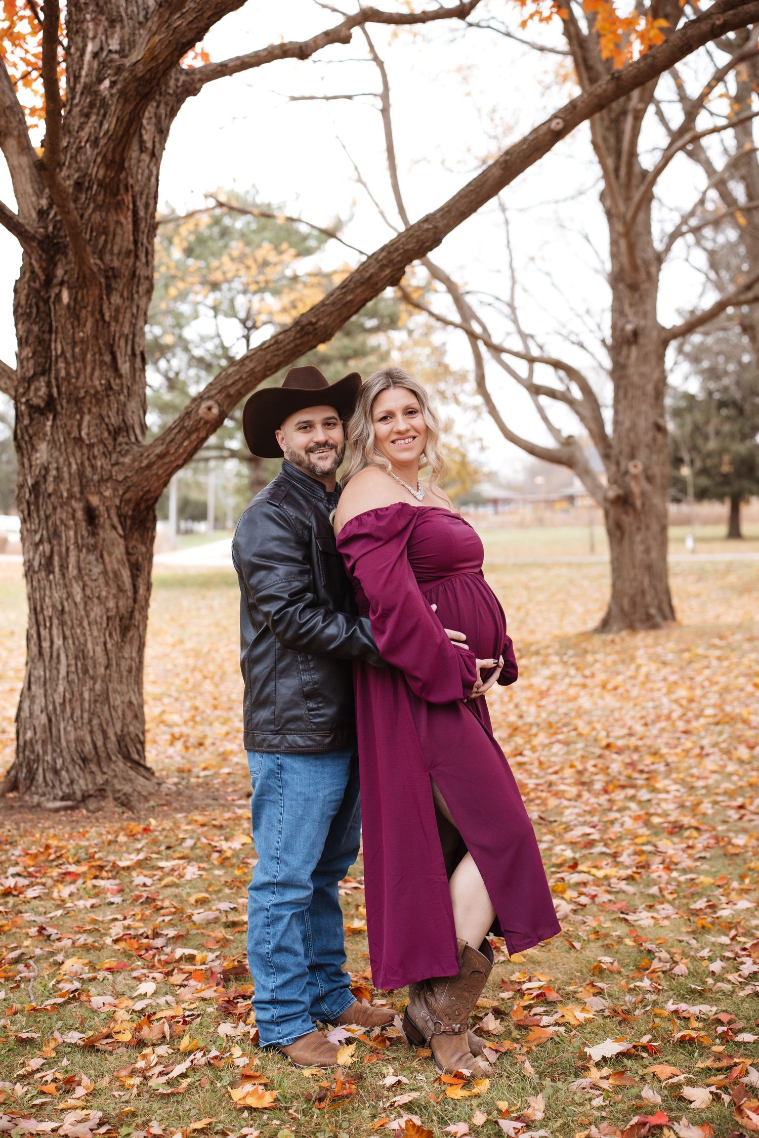 Couple in autumn park maternity shoot, man in cowboy hat and leather jacket embracing pregnant woman in burgundy dress.