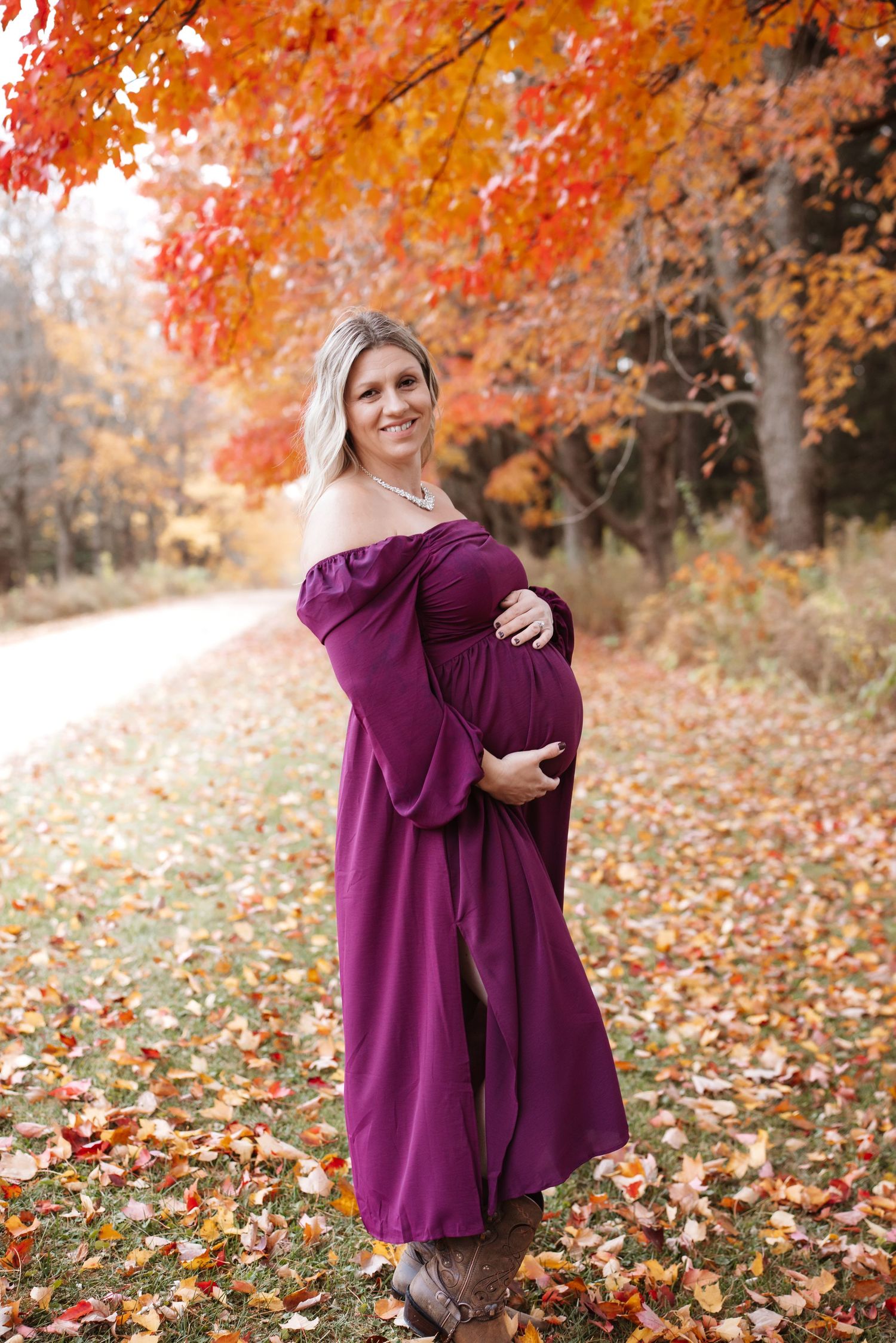 Pregnant woman in flowing purple off-shoulder dress standing on leaf-covered path in autumn park.
