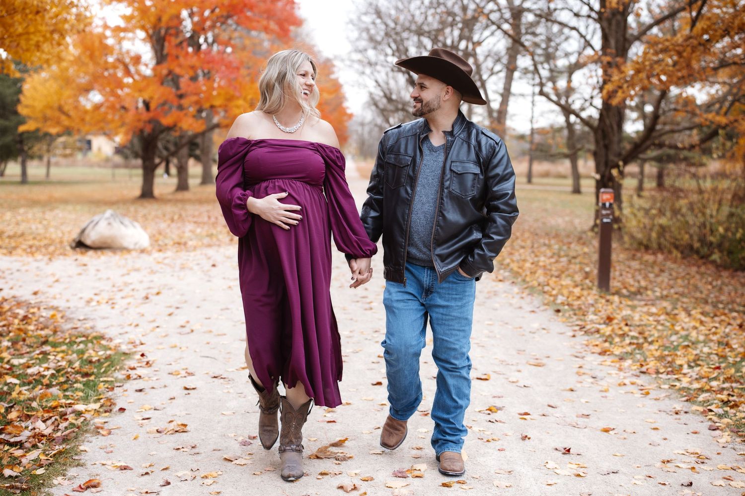 Pregnant couple walking hand-in-hand on a leaf-covered path surrounded by colorful autumn trees.