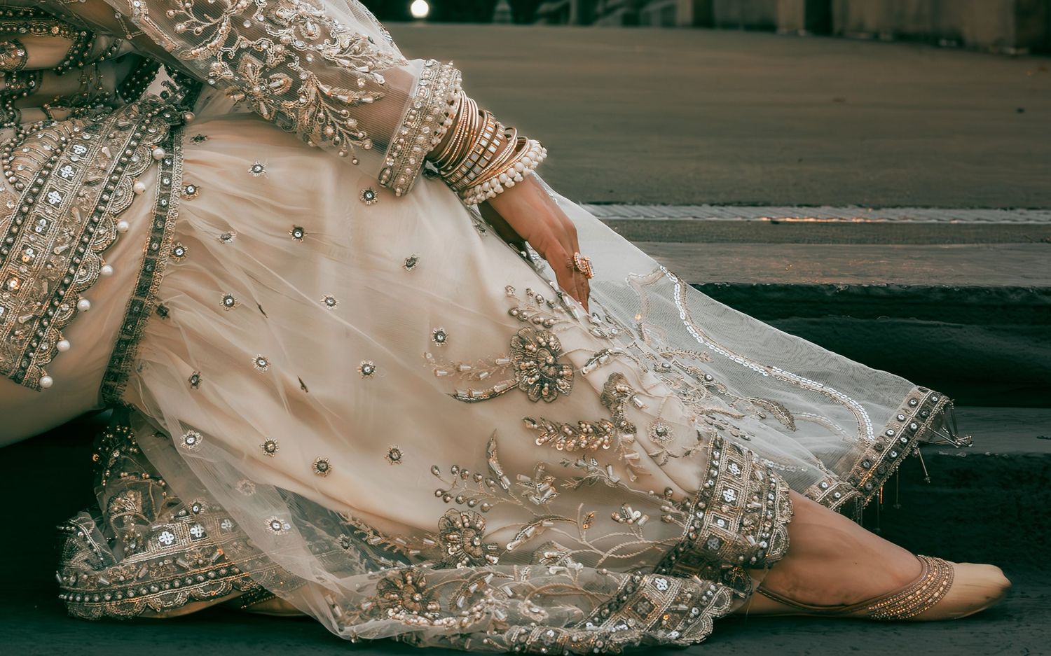 Close-up of South Asian bride's intricately embroidered ivory lehenga with gold bangles and jewelry.