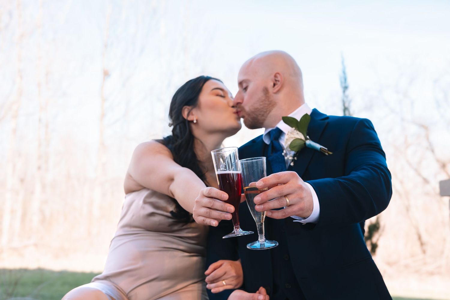 Newlyweds sharing a tender kiss while clinking red champagne glasses at their outdoor wedding reception.