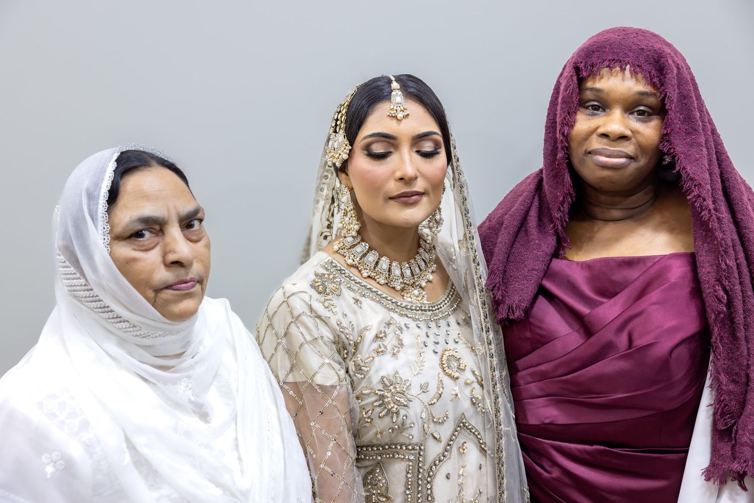 Islamic bride in gold embroidered white lehenga stands between two mothers in hijabs for wedding portrait.