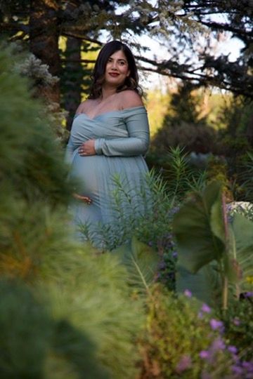 Expectant mother in mint green off-shoulder maternity dress standing amid colorful garden foliage and trees.