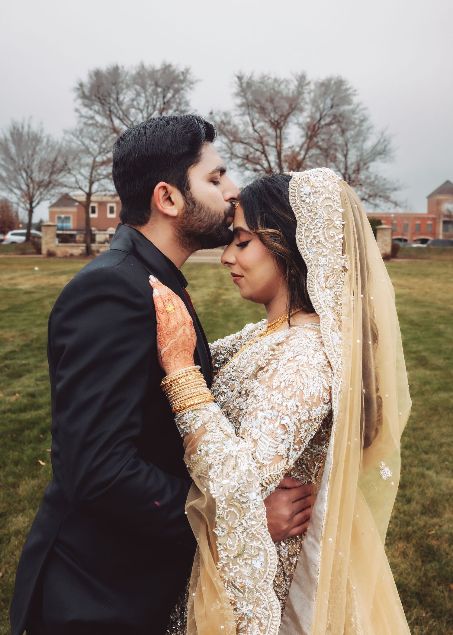 Groom in black suit kisses bride's forehead as she wears intricate white and gold bridal lehenga with veil.