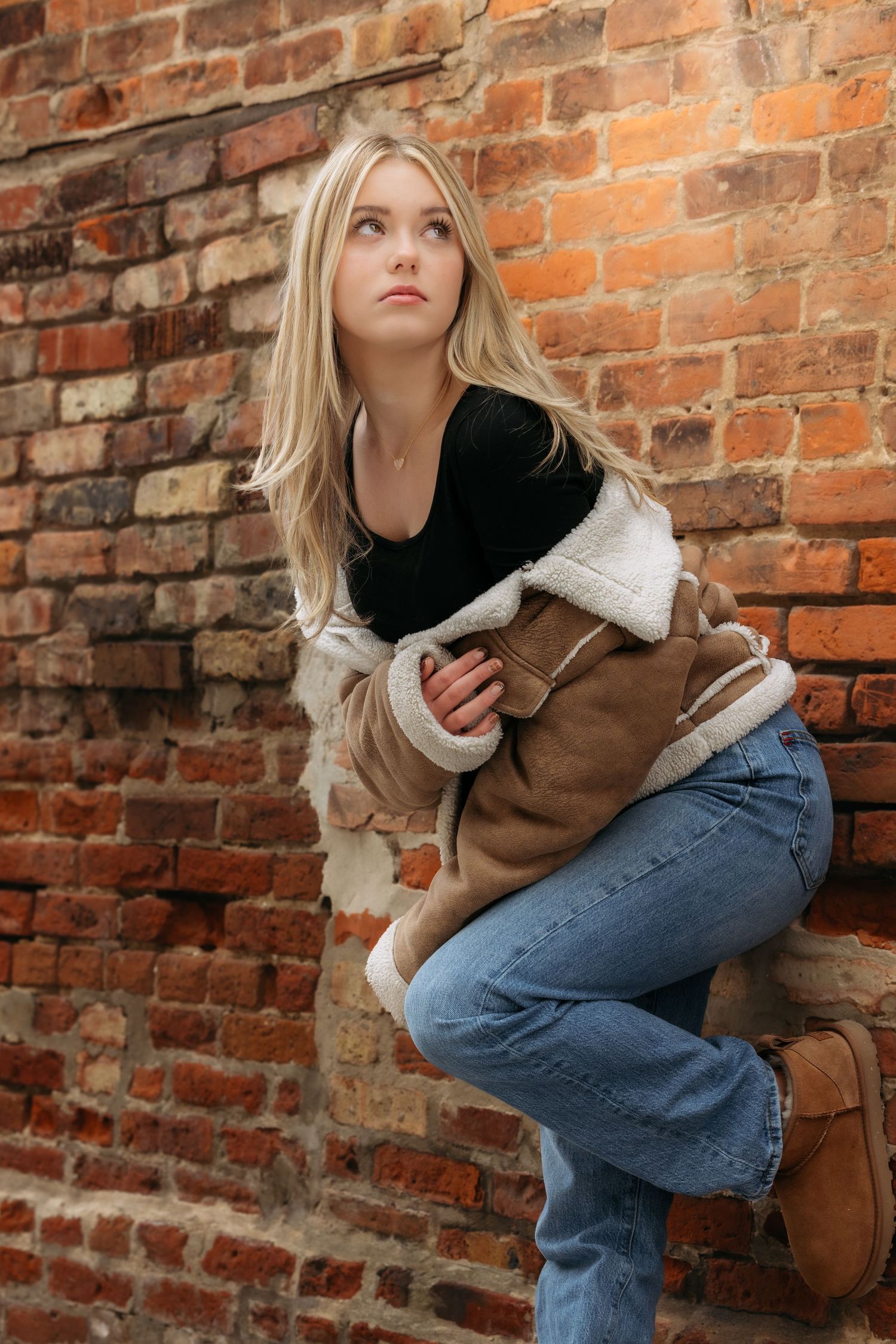A blonde woman in a shearling jacket and jeans sits against a rustic red brick wall looking upward.