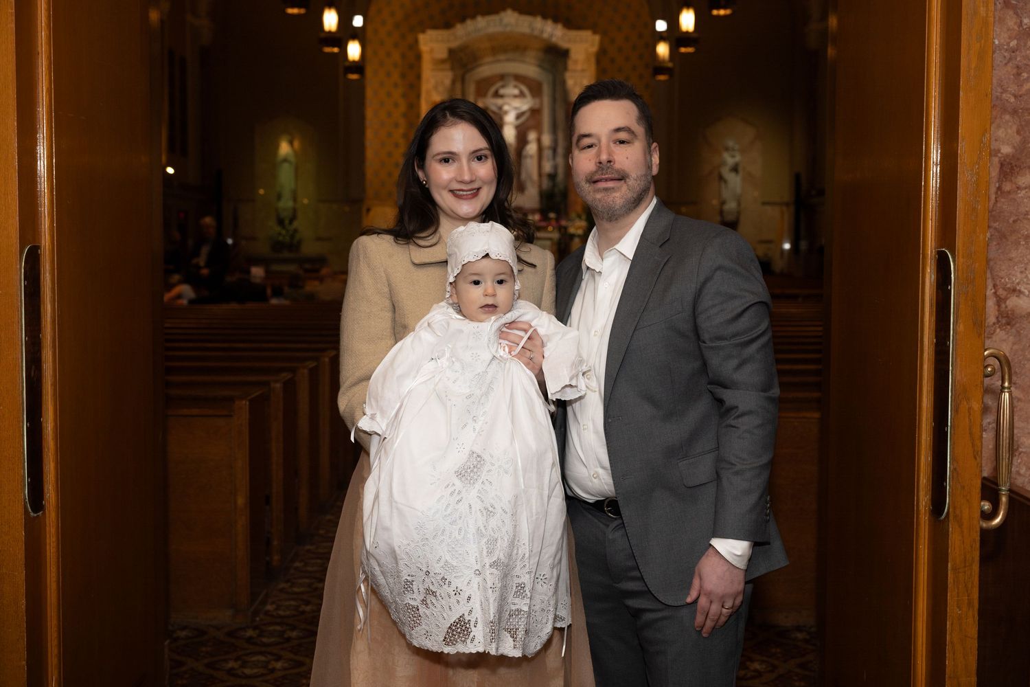 Couple in formal attire holding an infant in a white christening gown inside a Catholic church.