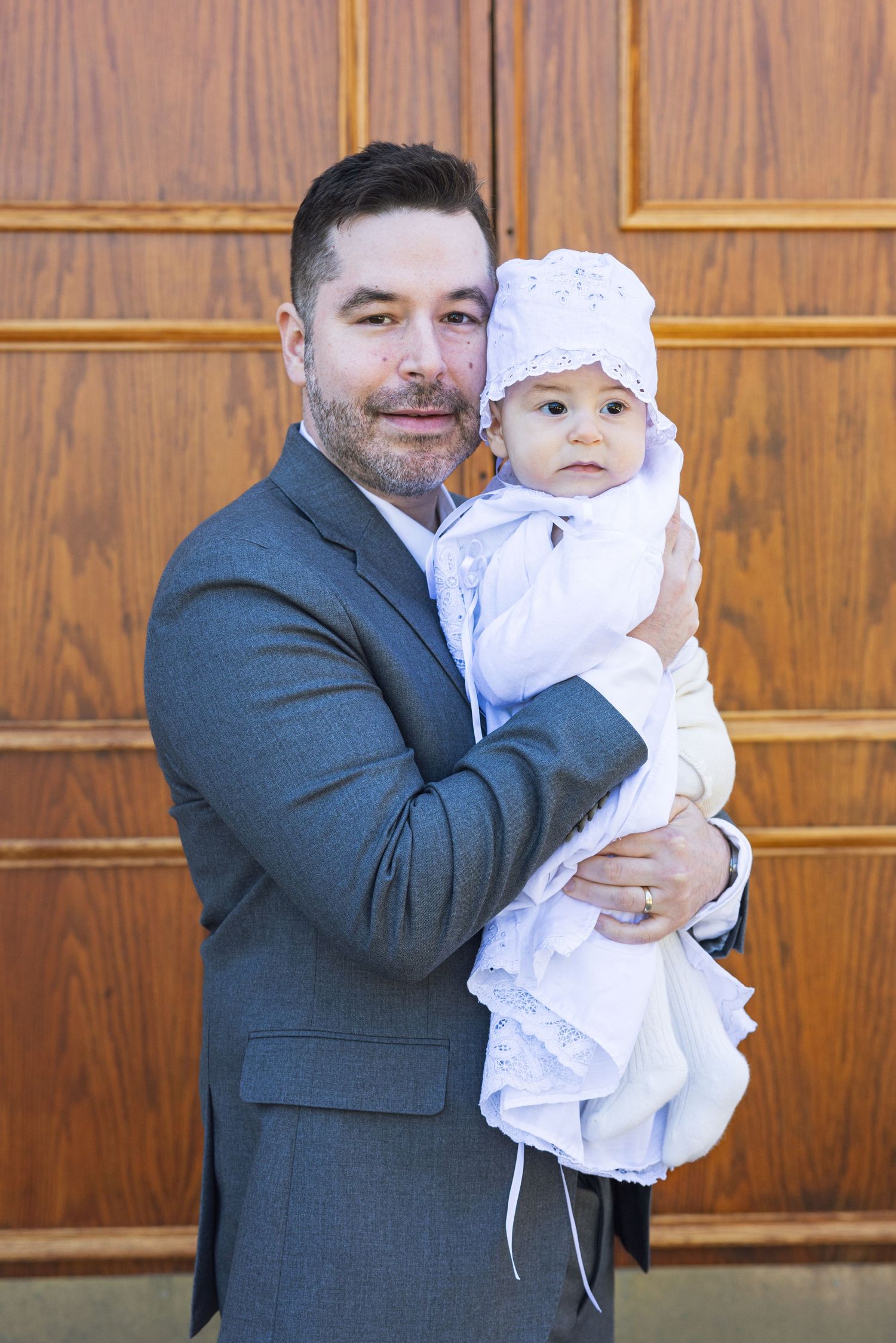Man in gray suit holding baby in white christening gown and bonnet at baptism ceremony.