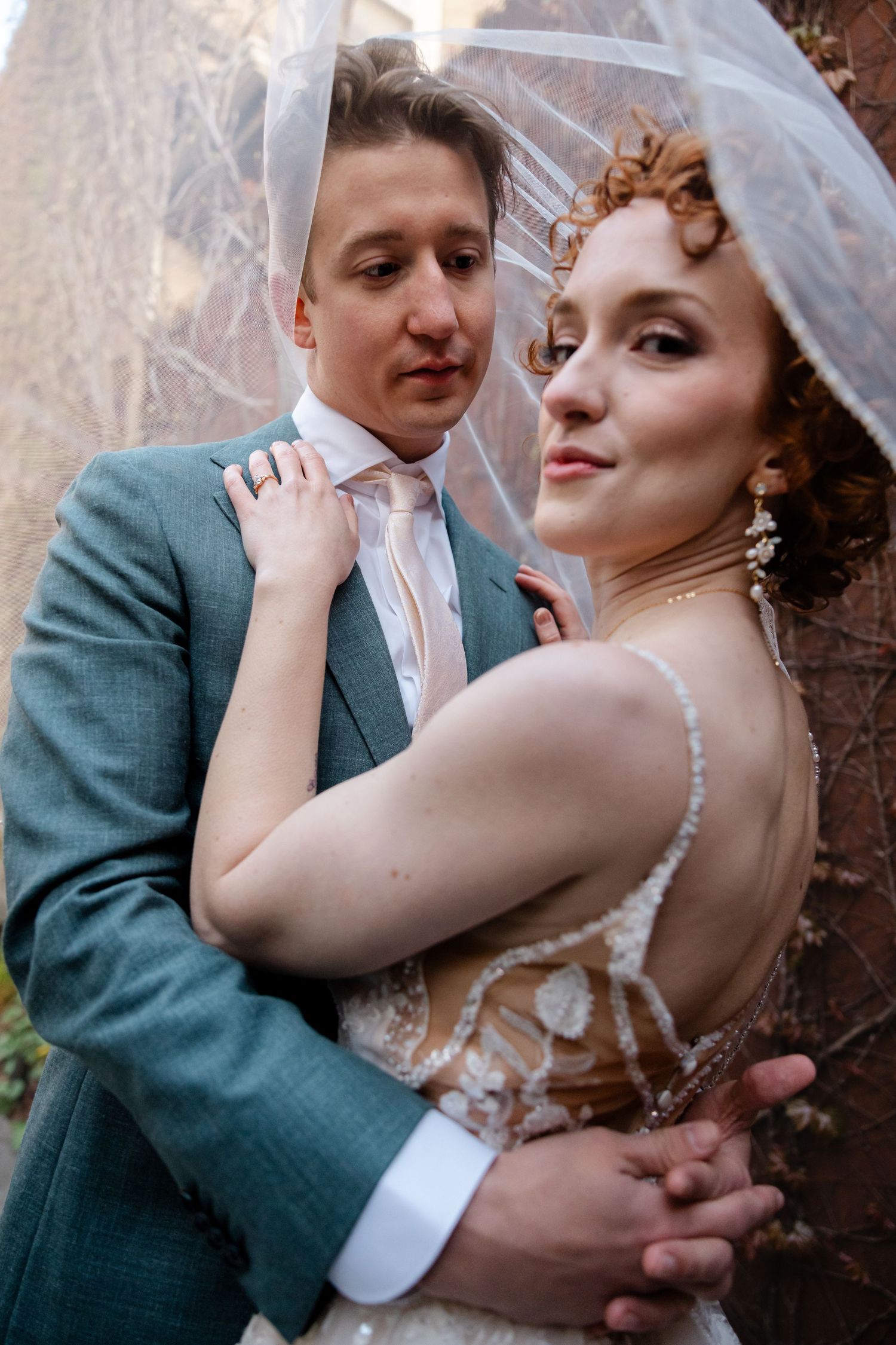 Groom in teal suit embraces bride in lace wedding dress as veil drapes over them both.