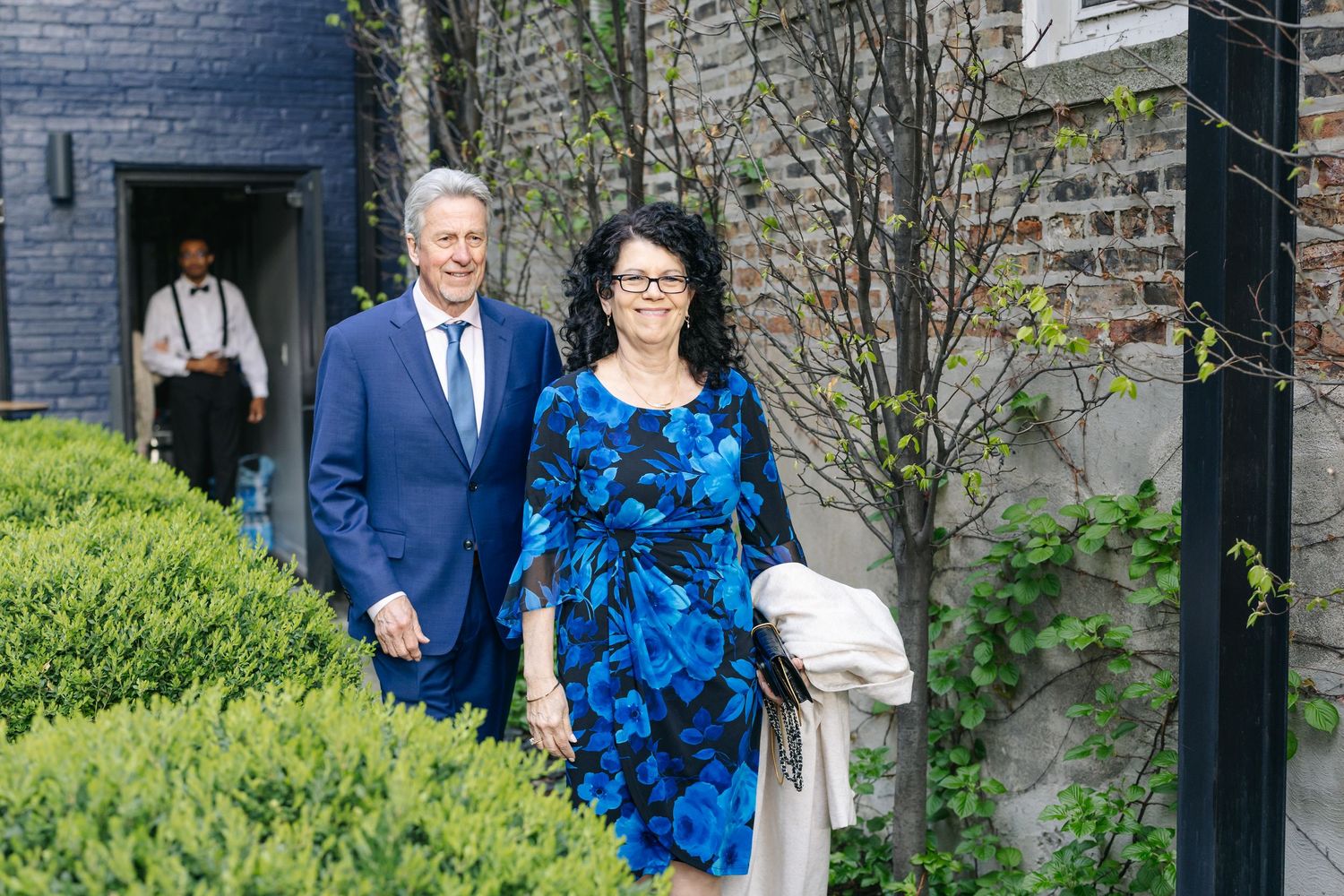 Elegant couple in blue attire walking outdoors, man in suit and woman in floral dress, at a formal garden event.