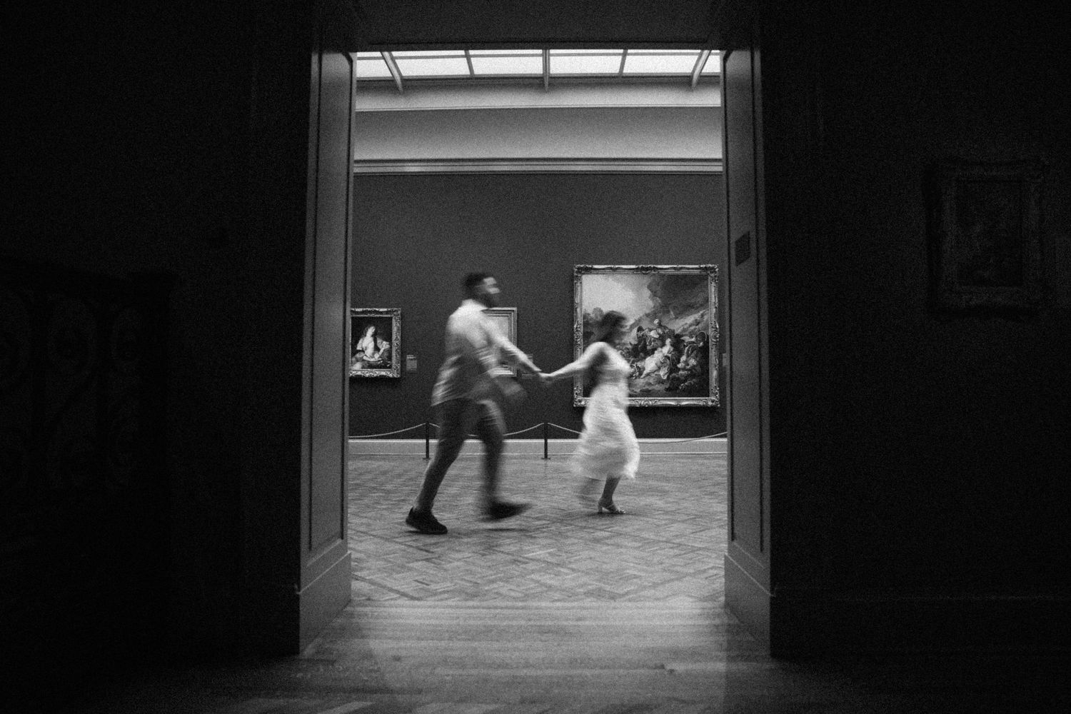 Black and white photo of couple running through grand museum gallery doorway during engagement shoot.